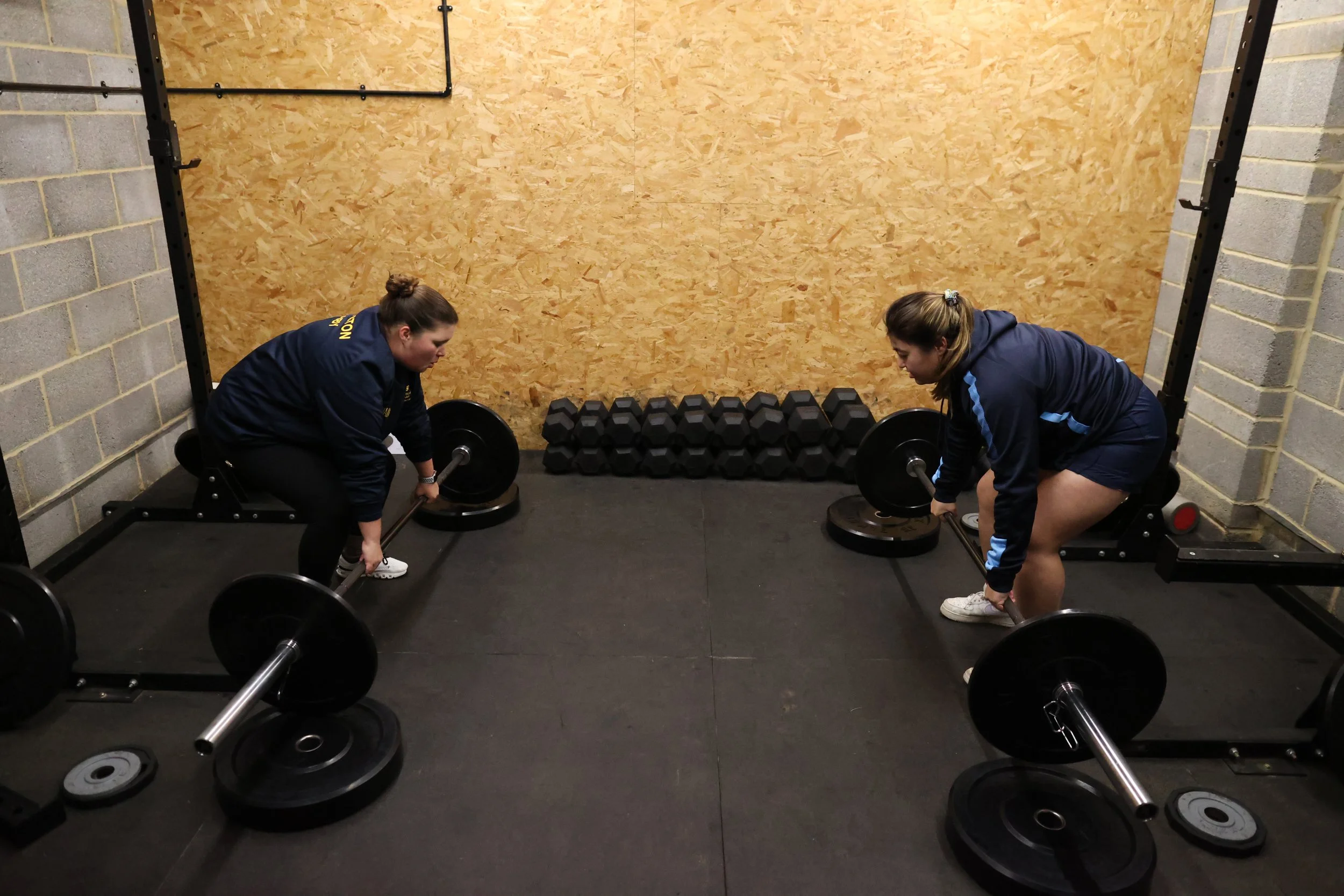 Two women in athletic clothing lifting barbells in a gym with black weight plates, a wooden wall, and a metal rack.