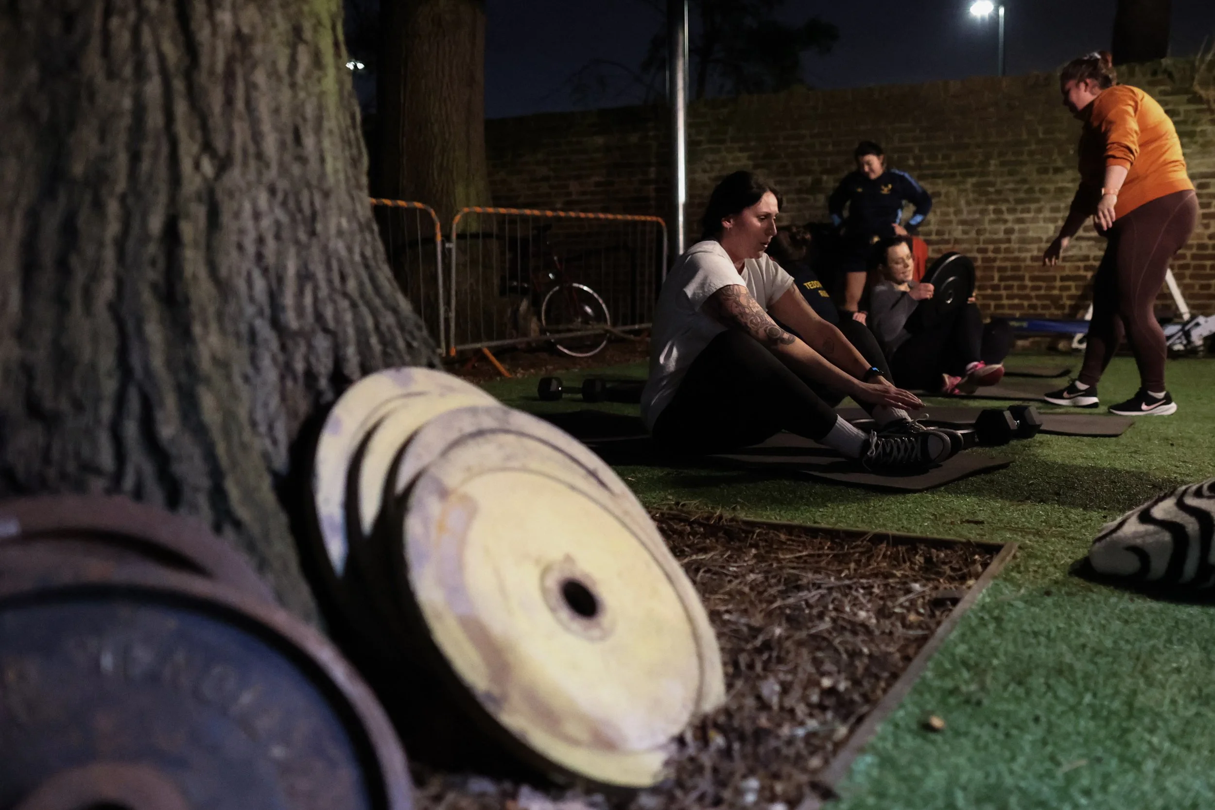 People exercising outdoors at night, sitting on exercise mats by a brick wall, with a tree in the foreground and bags nearby.
