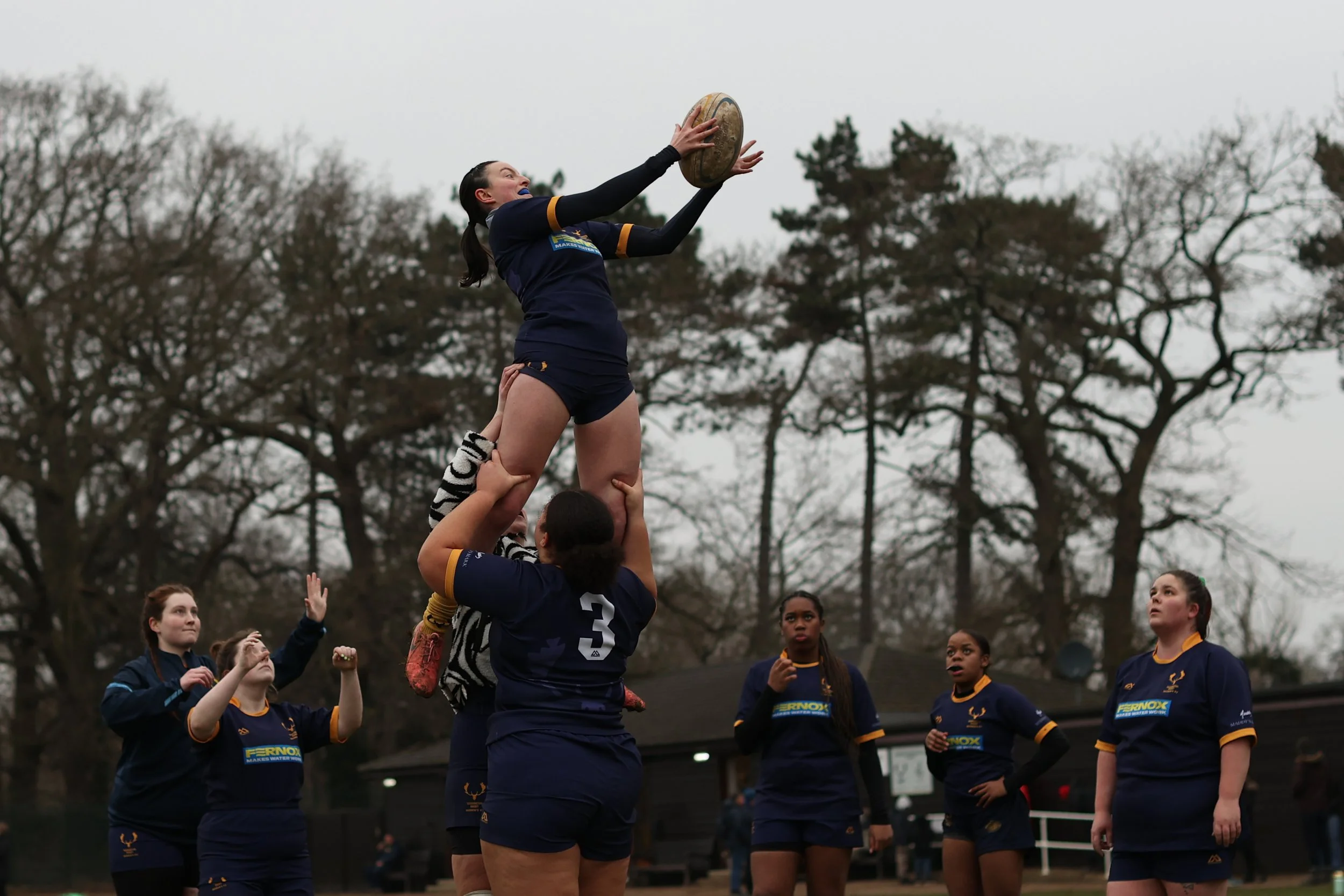 Women’s rugby team practicing lineout, with one player catching the ball as teammates lift her, outdoors during cloudy weather with trees in the background.