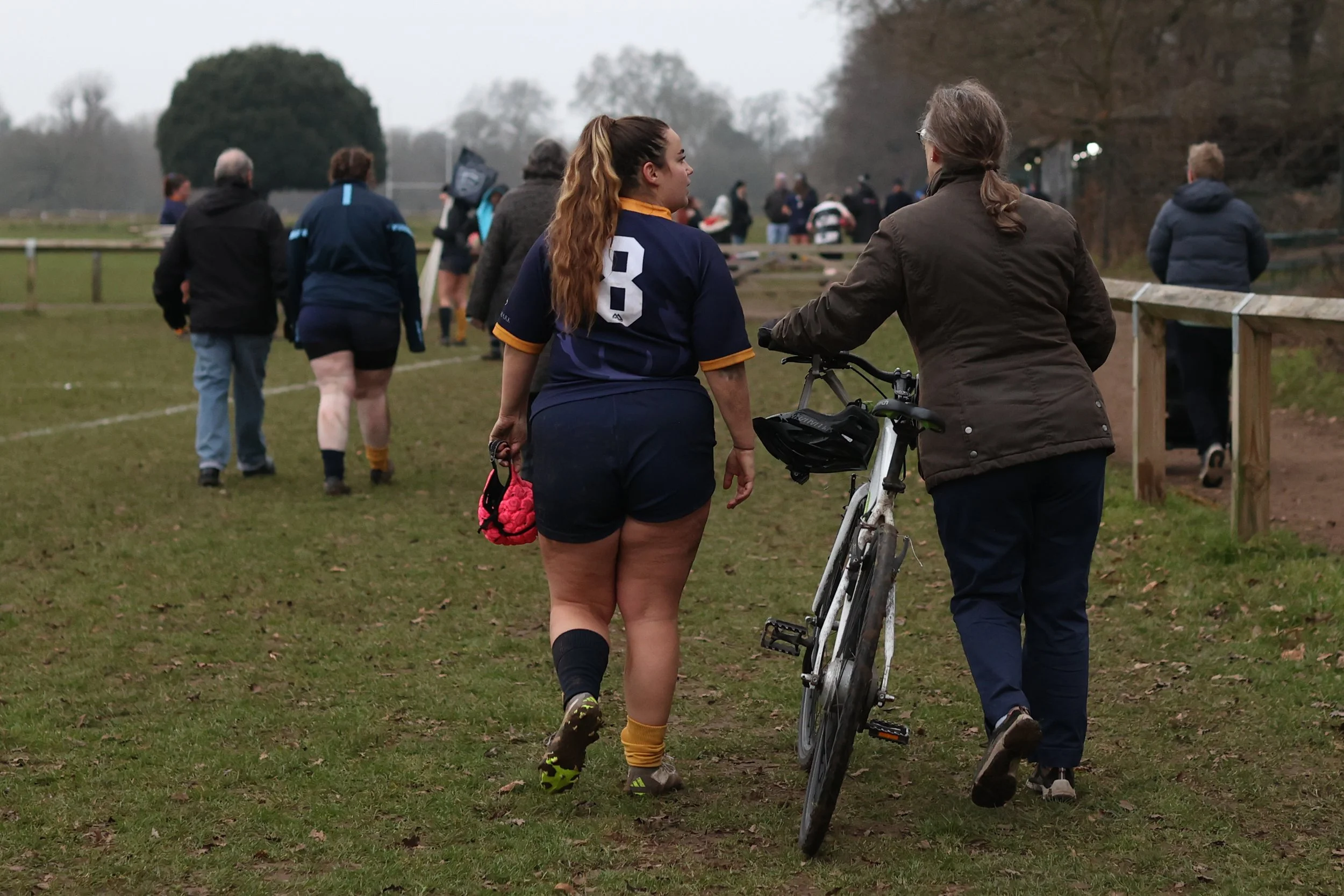 A group of people standing and walking on a grassy field, with two women in the foreground, one with a bicycle, and others in the background, dressed in sports and casual attire, on a cloudy day.
