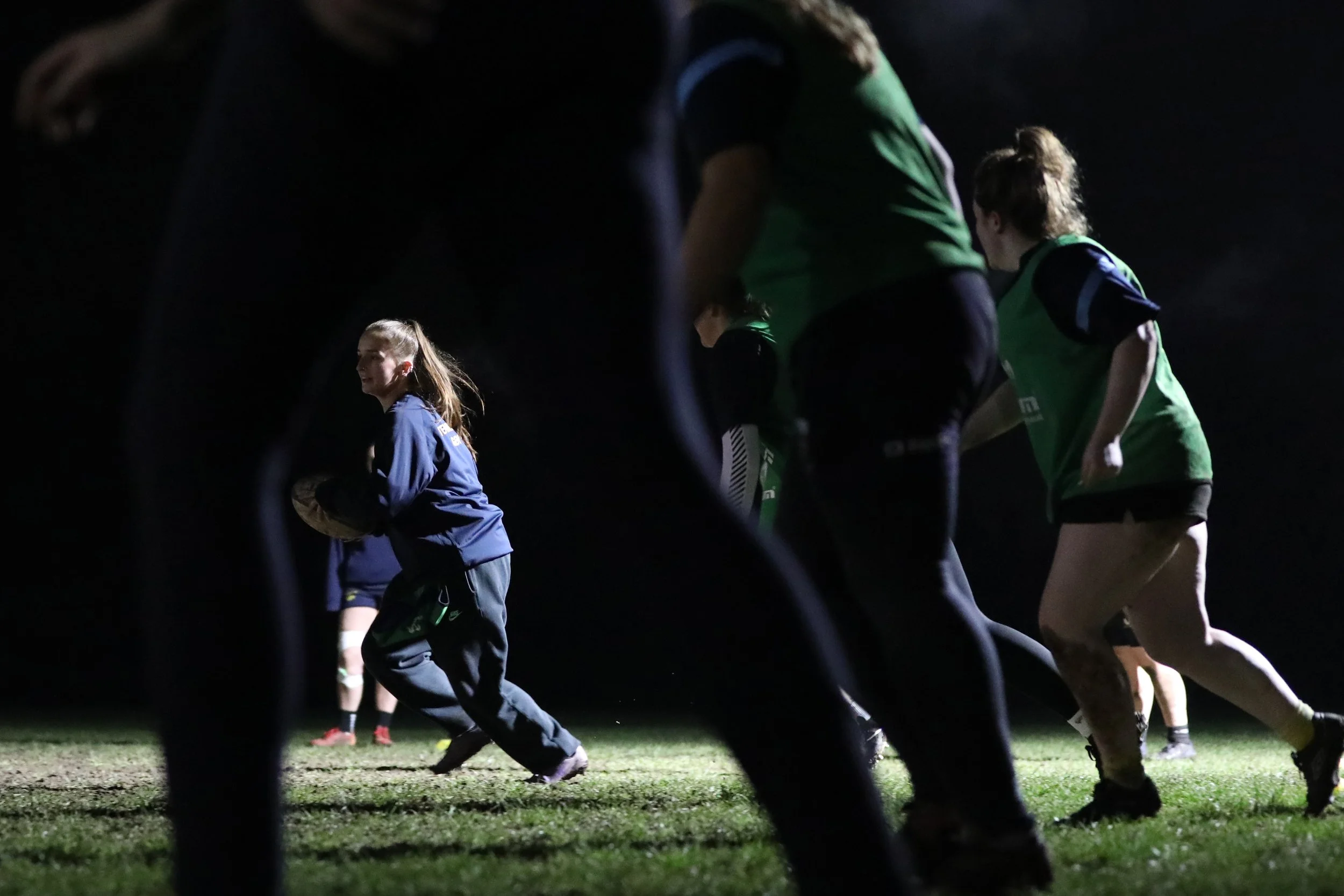 Nighttime soccer practice with young female athlete in blue uniform running on the field surrounded by players in green and black uniforms, seen through an opening in a fence.