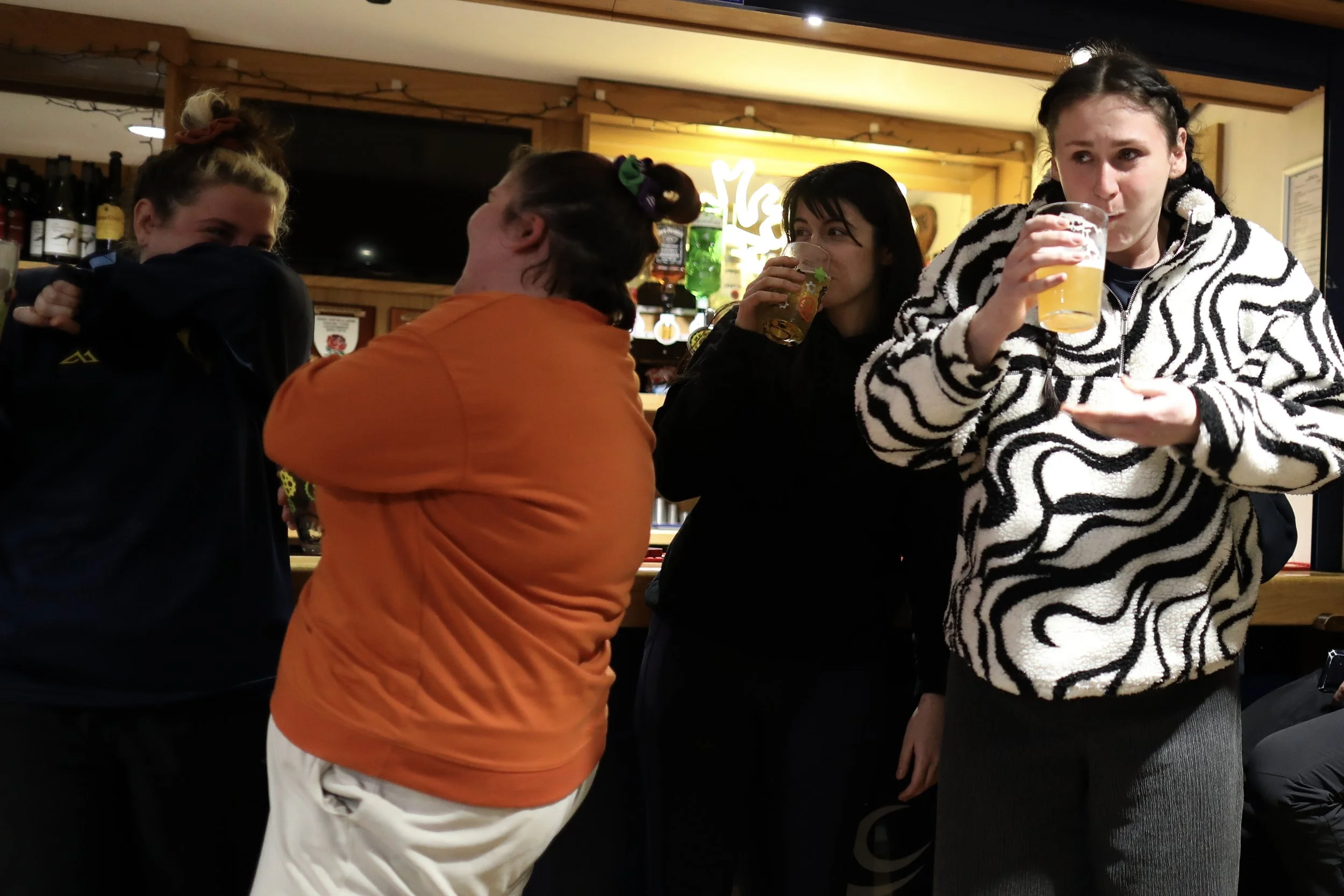Four women and one man are socializing at a bar, with three of them drinking beer from clear glasses. They are standing close together, engaged in conversation. The background shows a bar area with bottles and a neon sign.