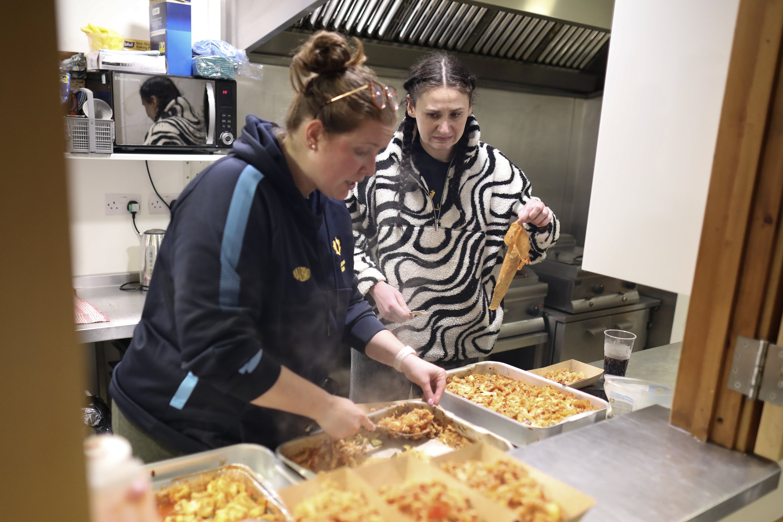 Two women serve themselves food from trays of pasta in a kitchen. One woman with her hair in a bun looks down at the food, while the other woman with braids holds a serving spoon and a napkin.
