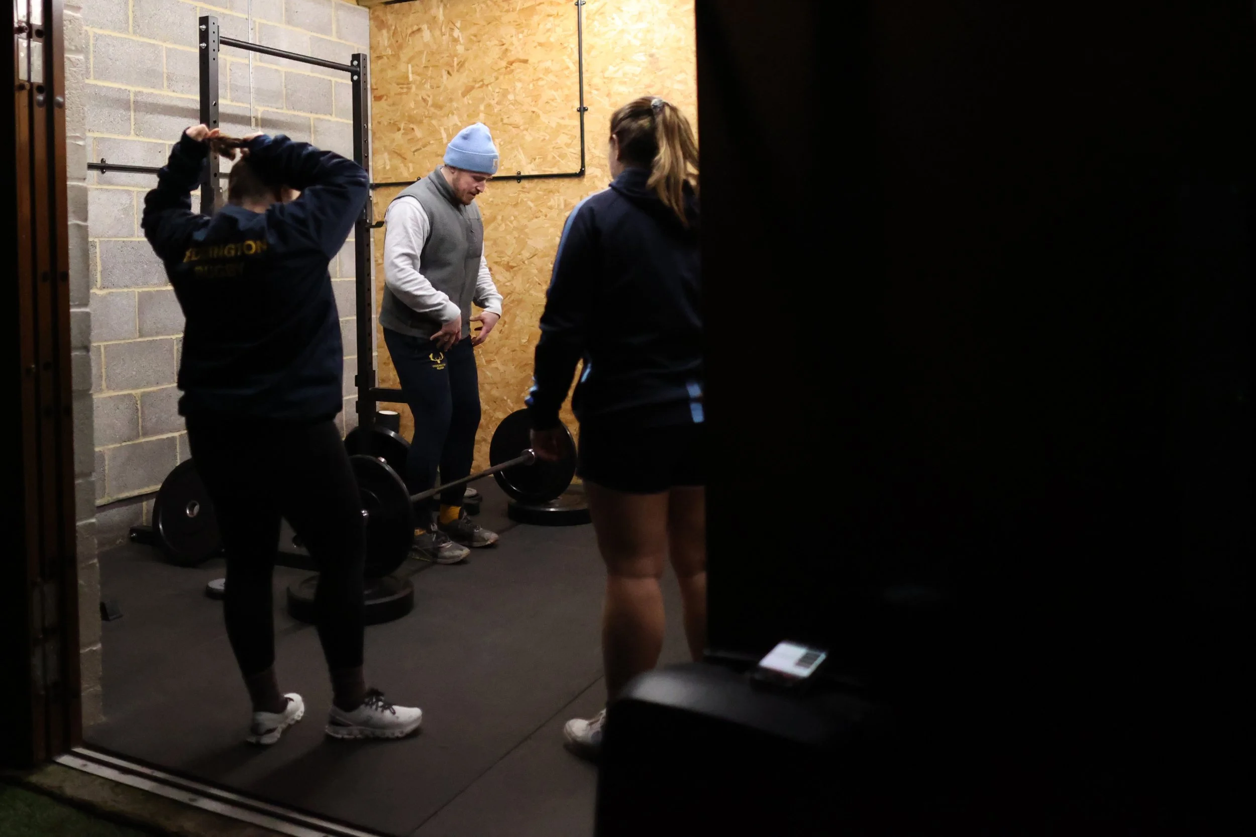 Group of three people, two women and one man, in a gym, preparing to lift barbells, with gym equipment and concrete walls in the background.