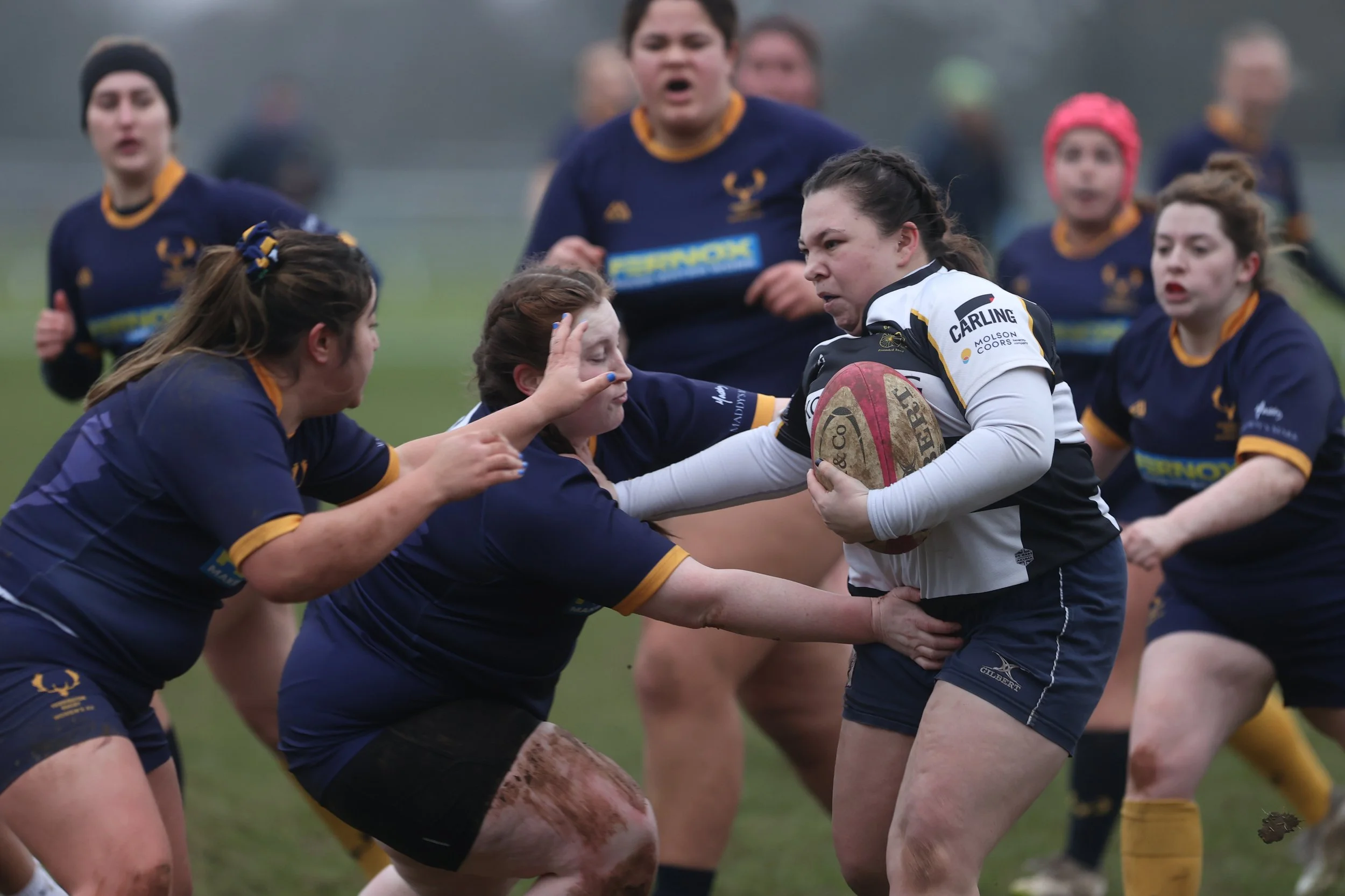 Women rugby players in a scrum during a match, wearing navy and white uniforms, on a grassy field.