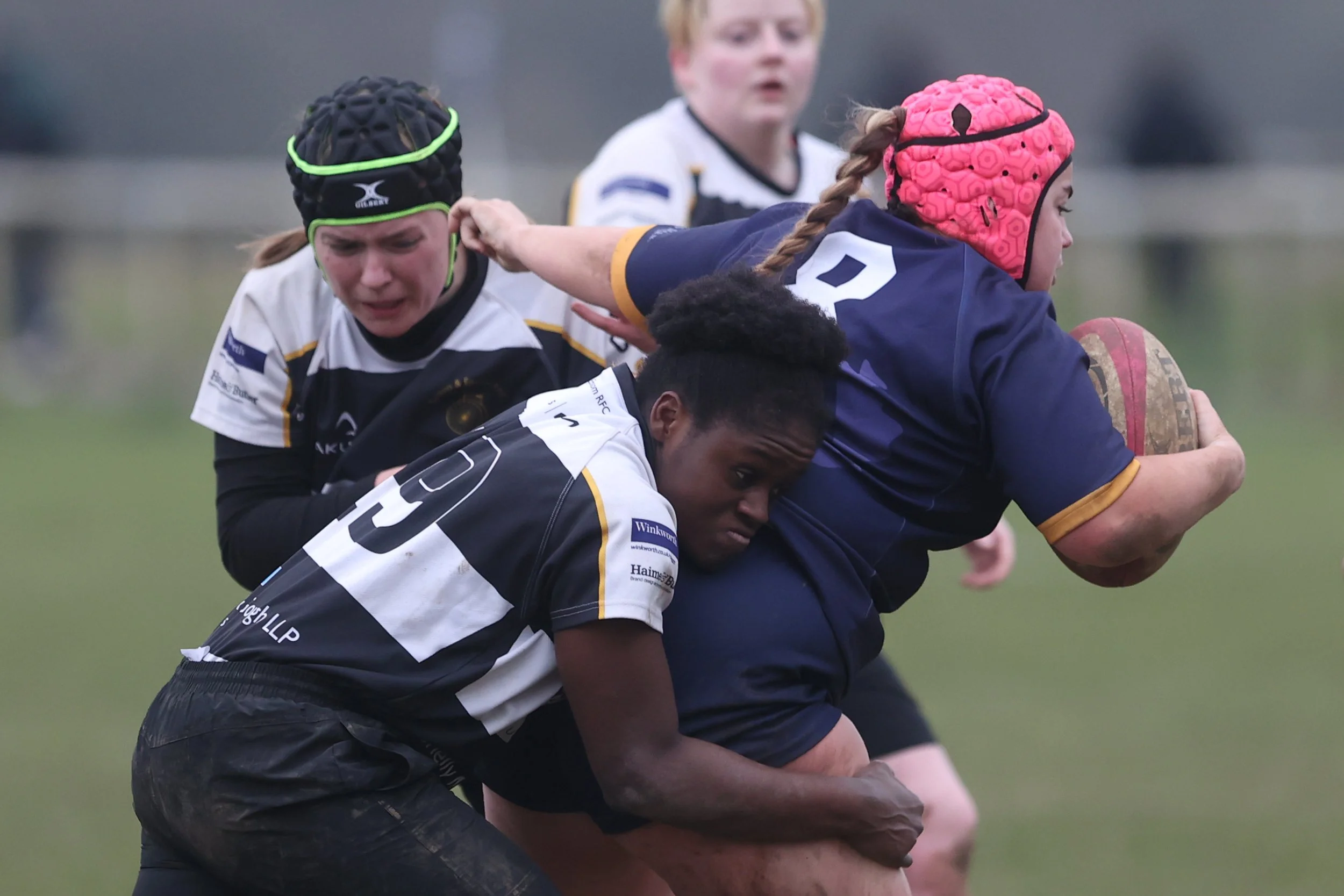 Women and girls playing rugby, tackling, and holding a rugby ball on a field during daytime.