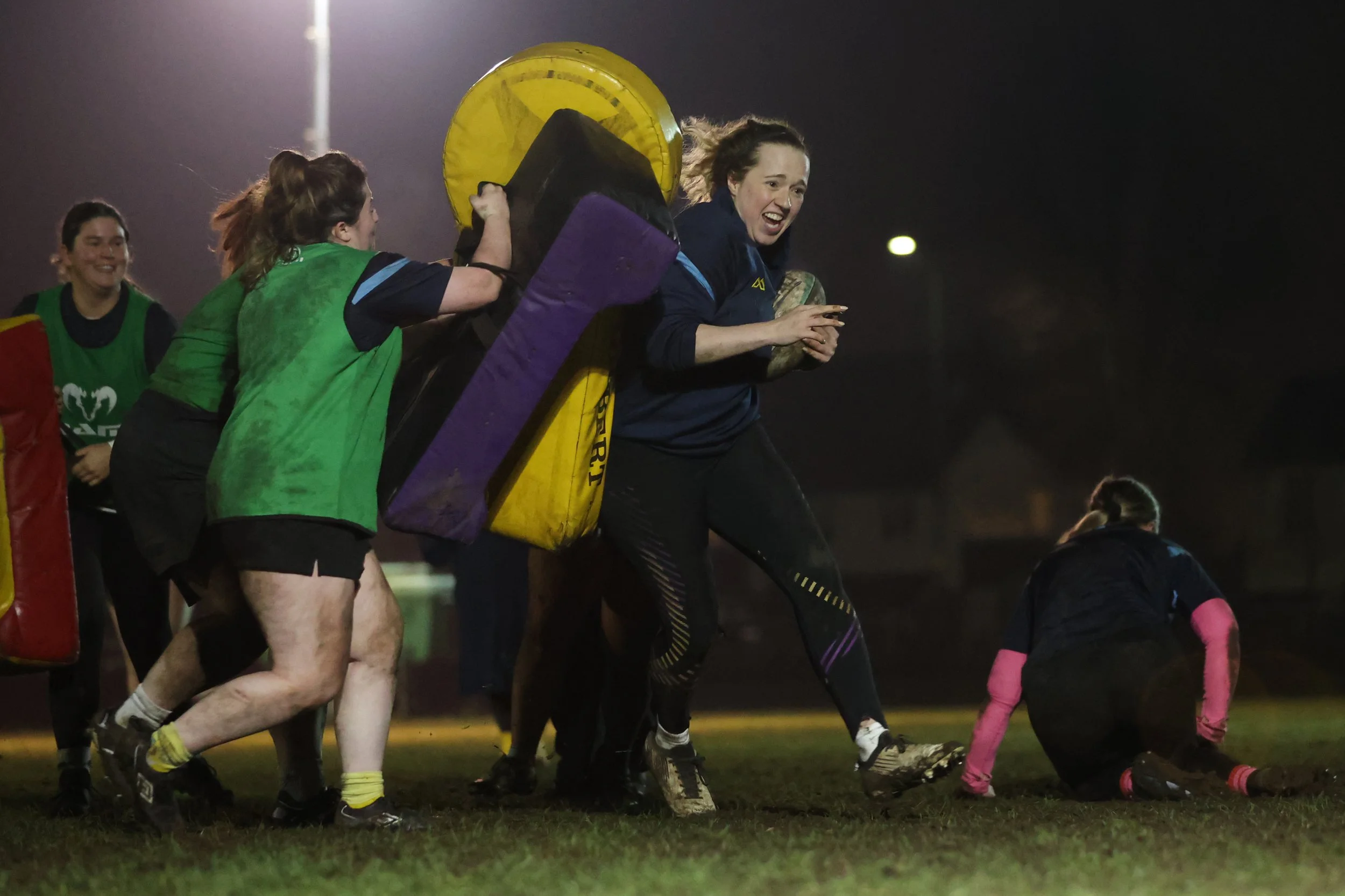 Women playing rugby at night, one woman falling while others help her up, on a grassy field under stadium lights.