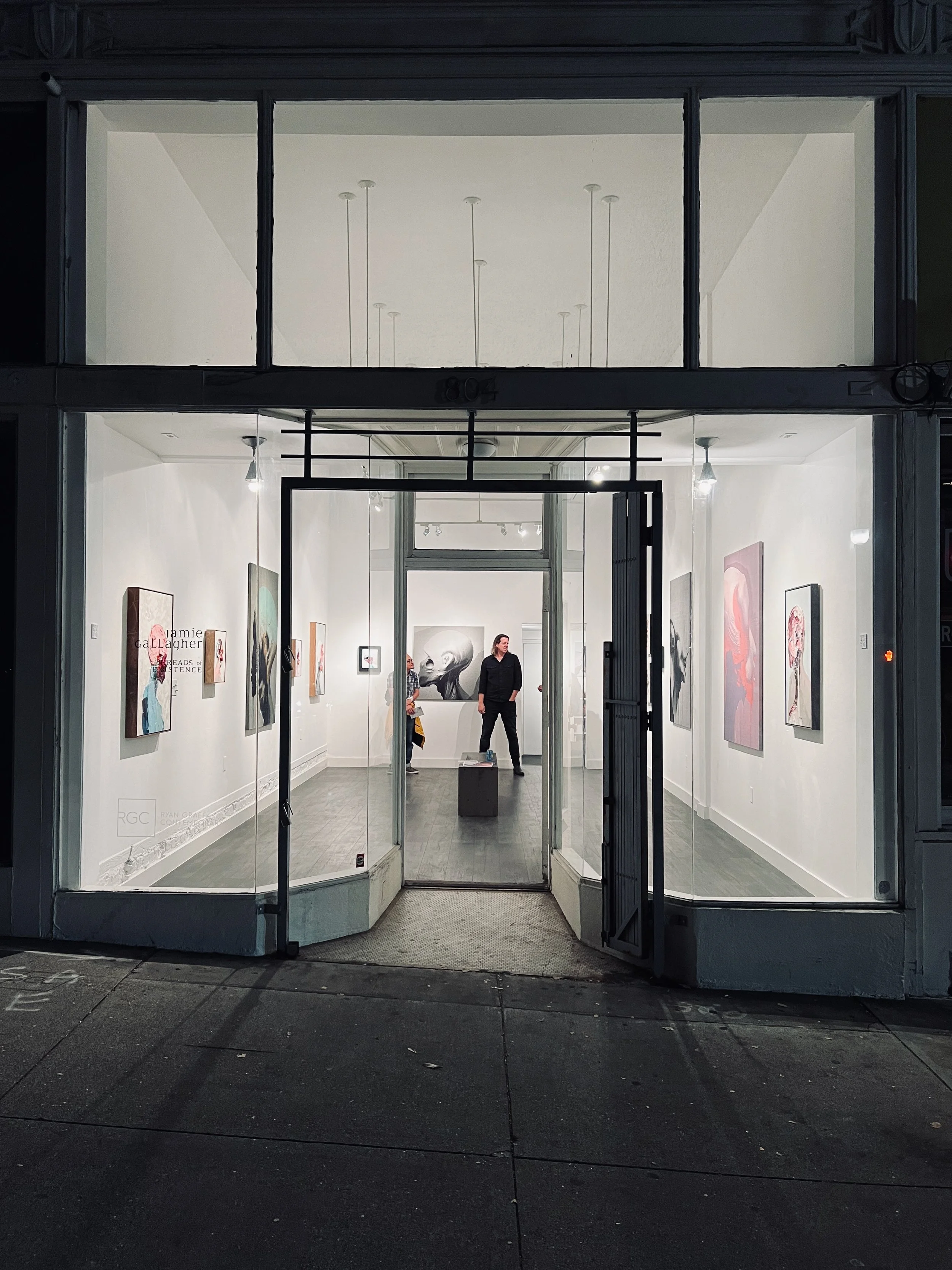 View through a large glass window of an art gallery interior with white walls, artwork on display, and a couple of people inside, one of whom is looking at the artwork while the other is standing near the back wall.