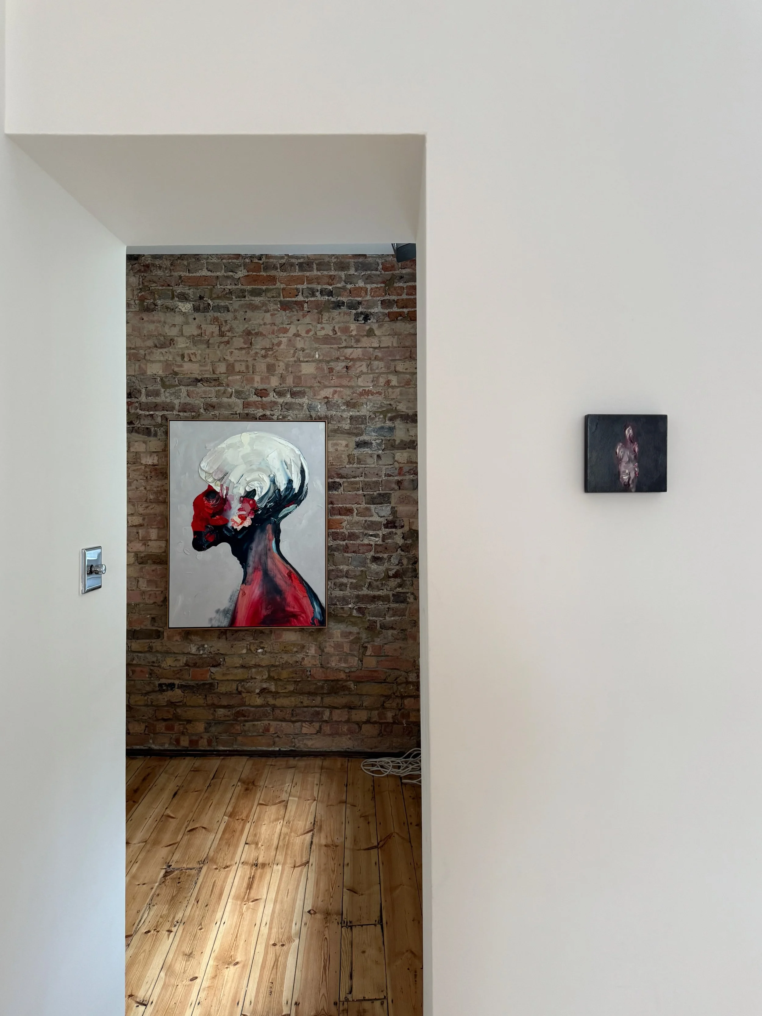 View of an art gallery with a brick wall displaying a portrait painting of a person with a white head and a red and black face, and a small dark abstract painting on a white wall to the right.