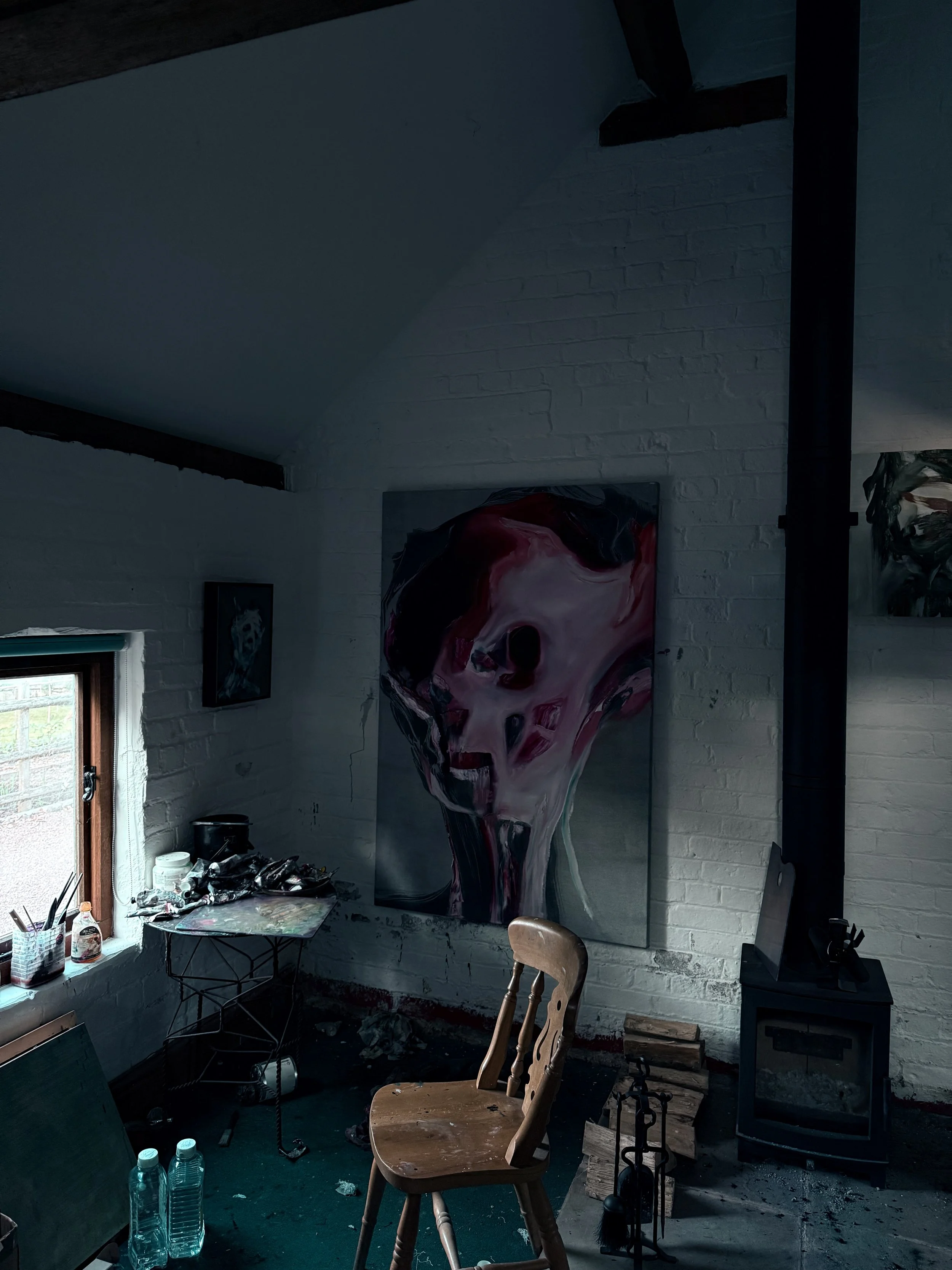 Artist's studio with a large abstract painting of a skull on the brick wall, a wooden chair, art supplies on a table, and a window letting in natural light.