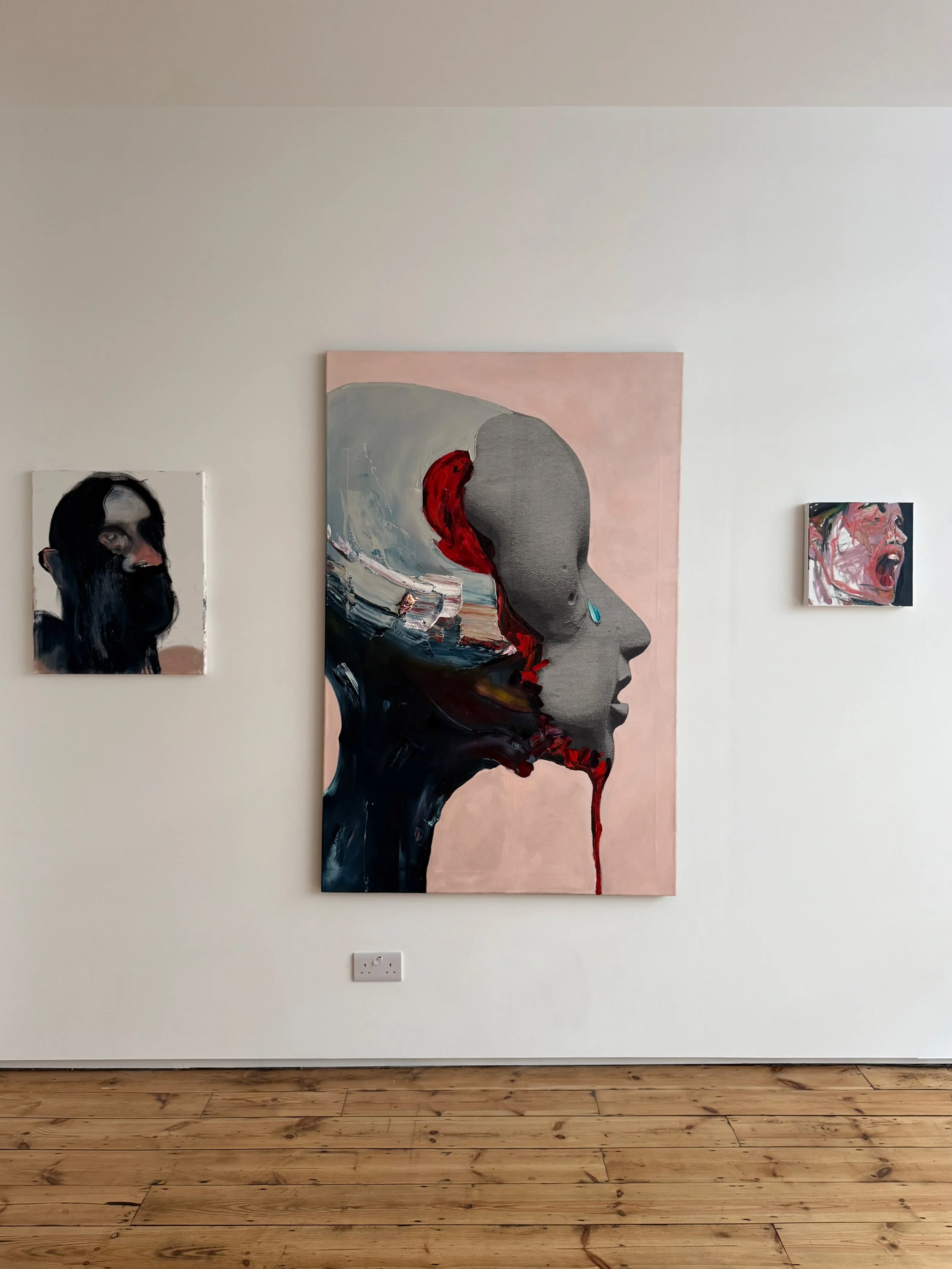 Art gallery wall with three paintings: a large abstract portrait of a face in profile with a tear, a small portrait of a person with long dark hair, and a small expressive face with wild hair.
