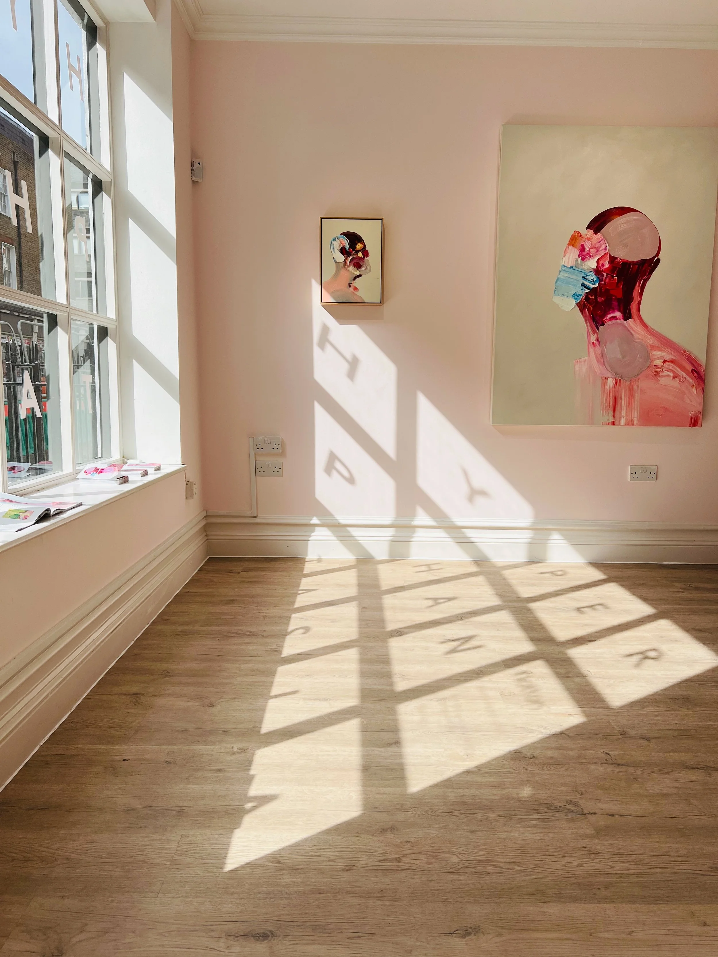 Sunlight creates shadow of window frame and letters on a light wood floor in a gallery with pastel pink walls, displaying two painted portraits of women with face masks.
