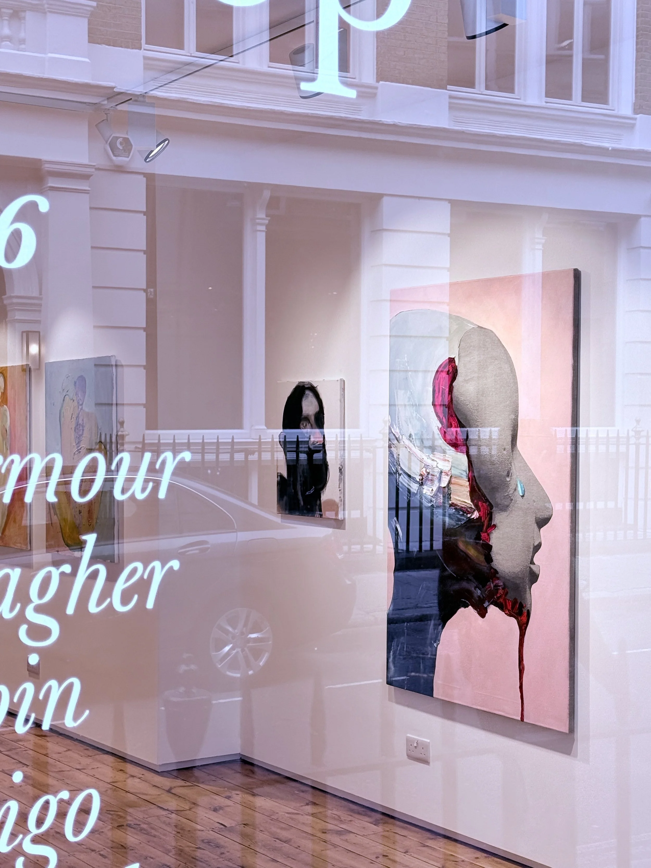 Interior view of an art gallery with paintings on the wall, including a portrait of a woman with a split face and abstract art, seen through a glass window with reflection of the street outside.