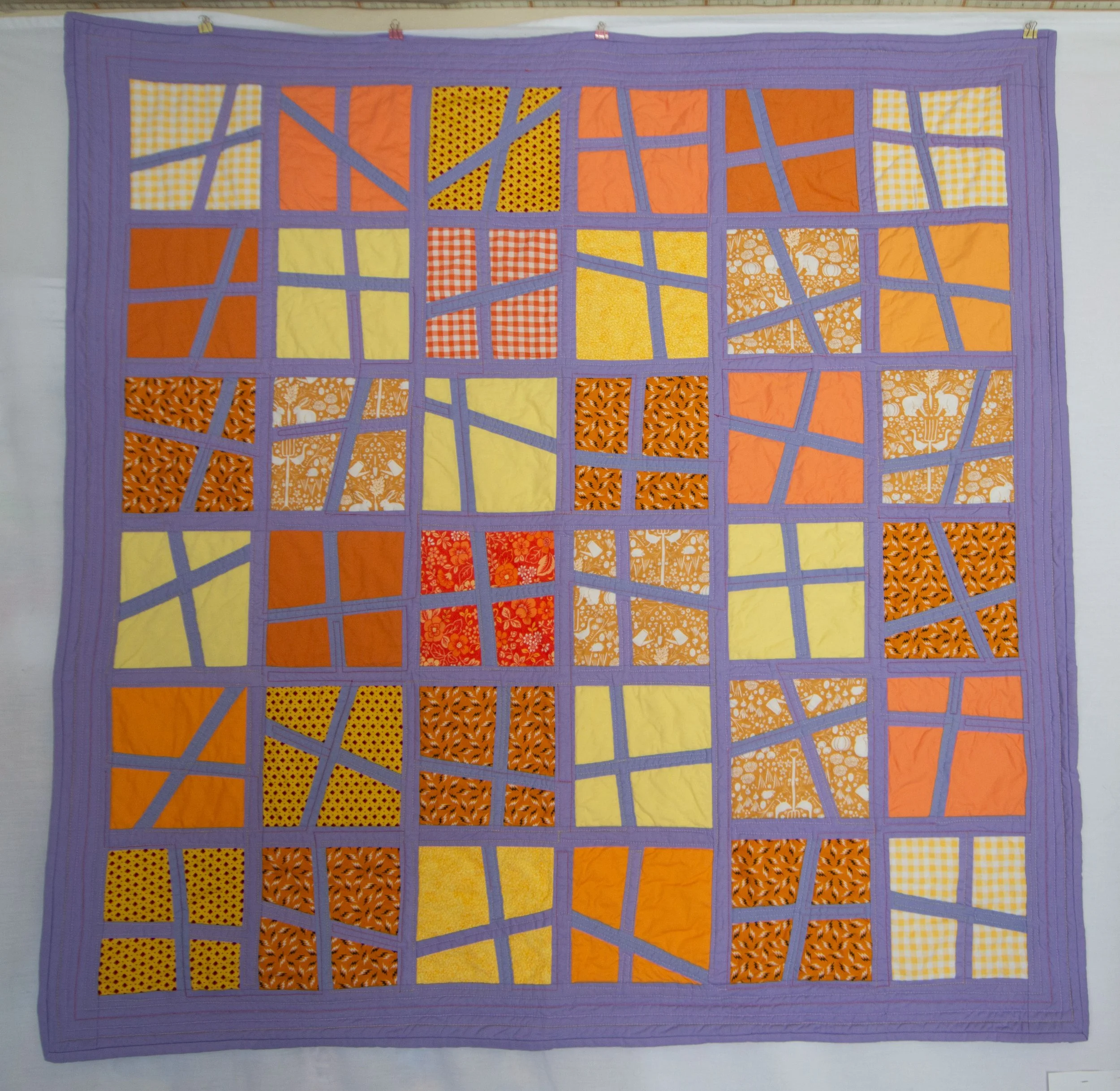 A quilt with an abstract city map design, featuring various orange, yellow, and patterned fabric patches outlined with purple border strips.