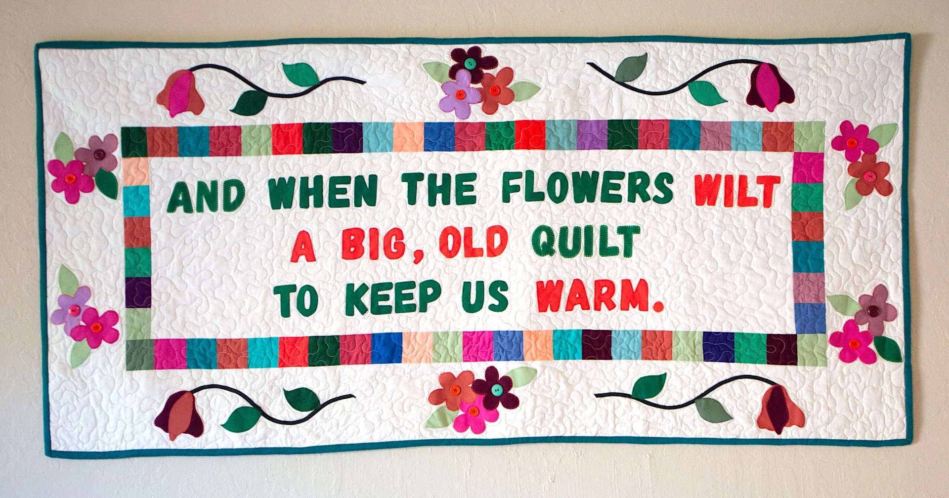 Colorful quilt wall hanging with flowers and a quote about flowers and warmth.
