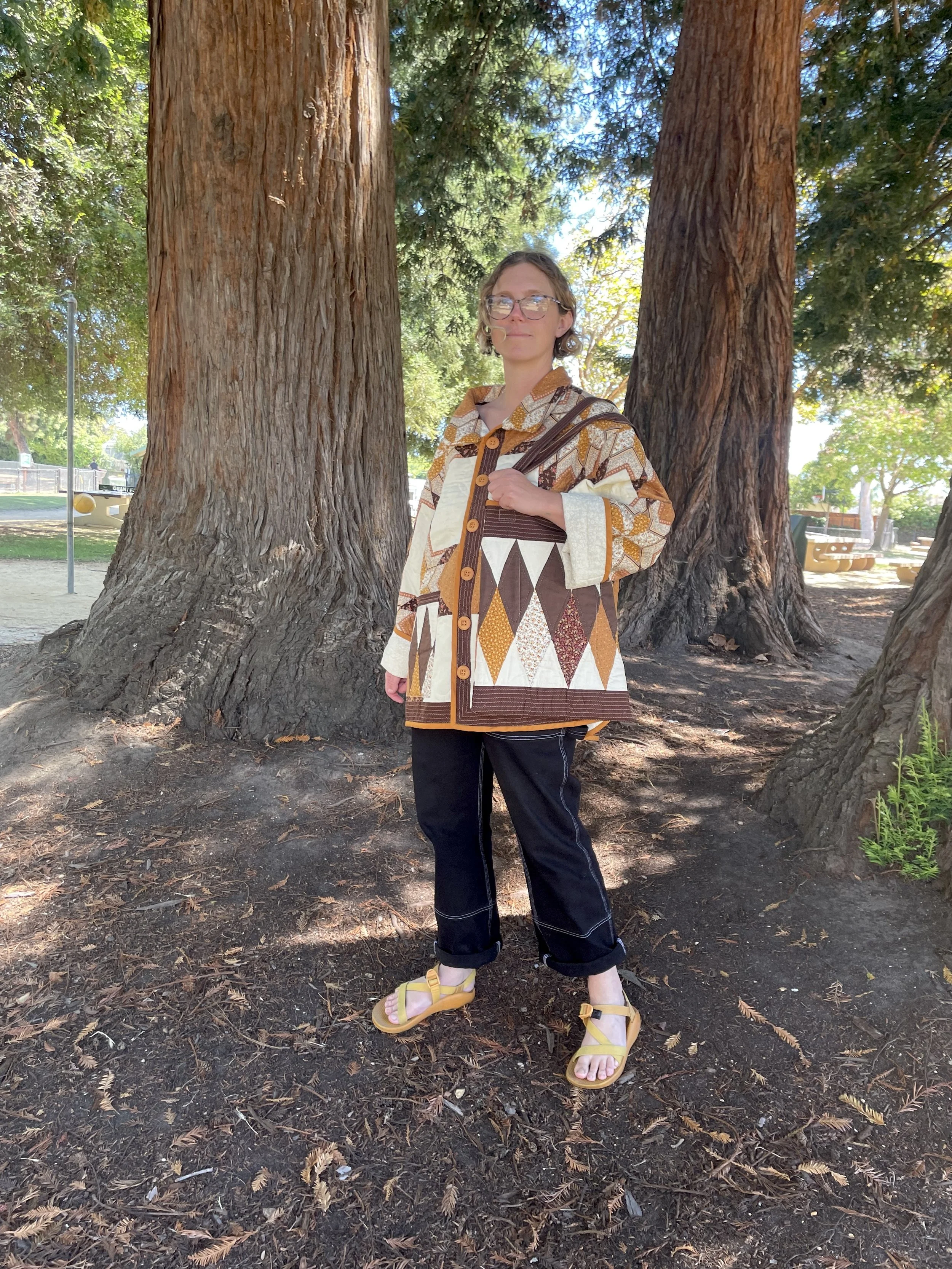 woman standing among redwood trees wearing a brown quilt coat and quilted bag