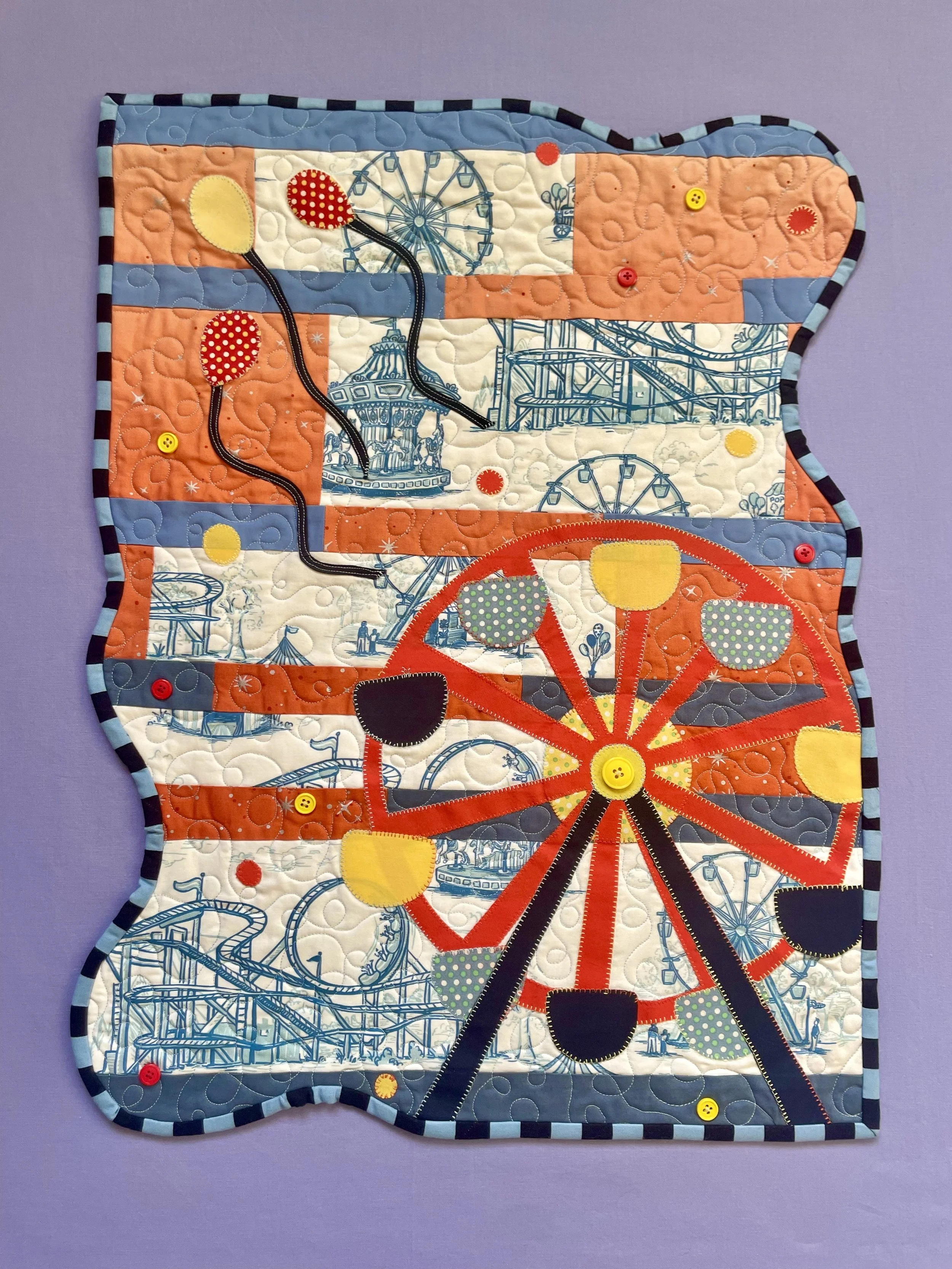 A quilted fabric piece featuring a carnival amusement park scene, including a Ferris wheel, roller coaster, and carousel. The quilt has colorful stitching, buttons, and a border with blue and black stripes, set against a purple background.