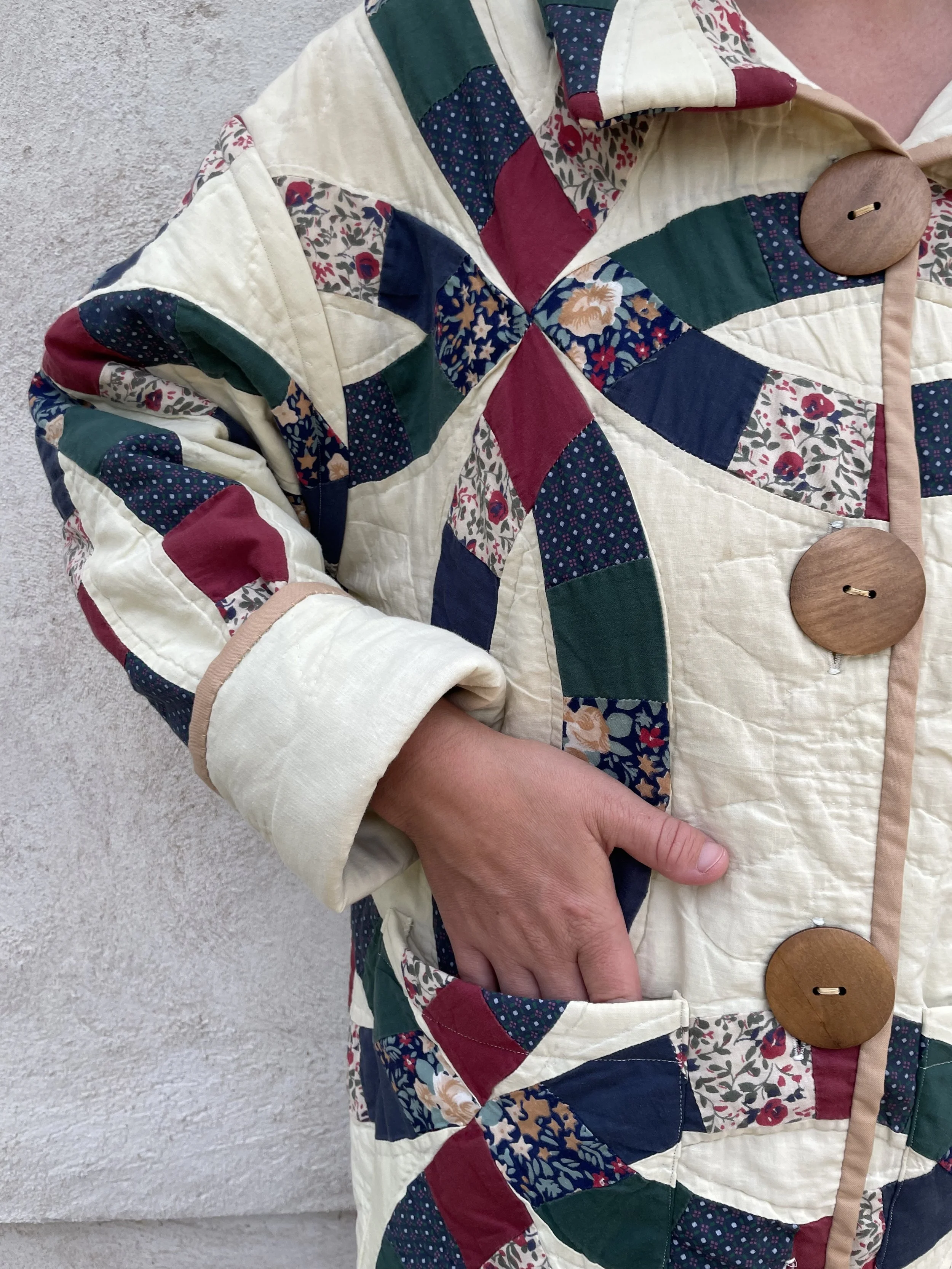 A person wearing a quilted jacket with a patchwork pattern of various colorful floral and geometric fabrics, with large wooden buttons and beige trim, standing with one hand in their pocket against a plain textured wall.