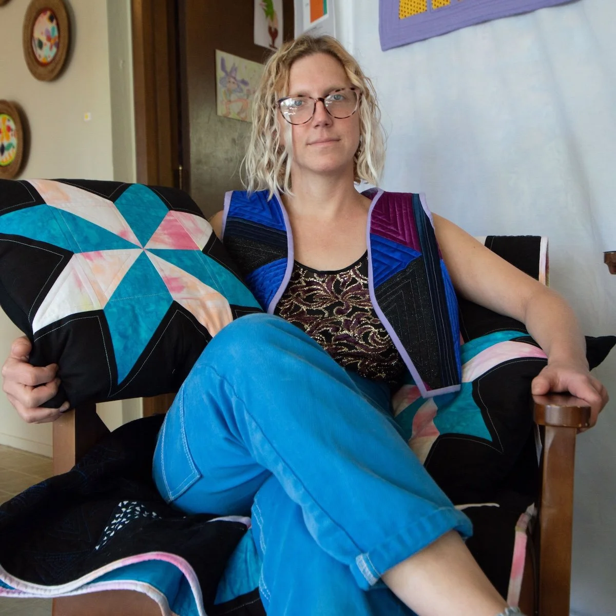 A woman with curly blonde hair and glasses sitting on a wooden chair, holding a colorful quilted pillow, wearing a sleeveless patterned top and blue pants, in a vibrant, decorated room.