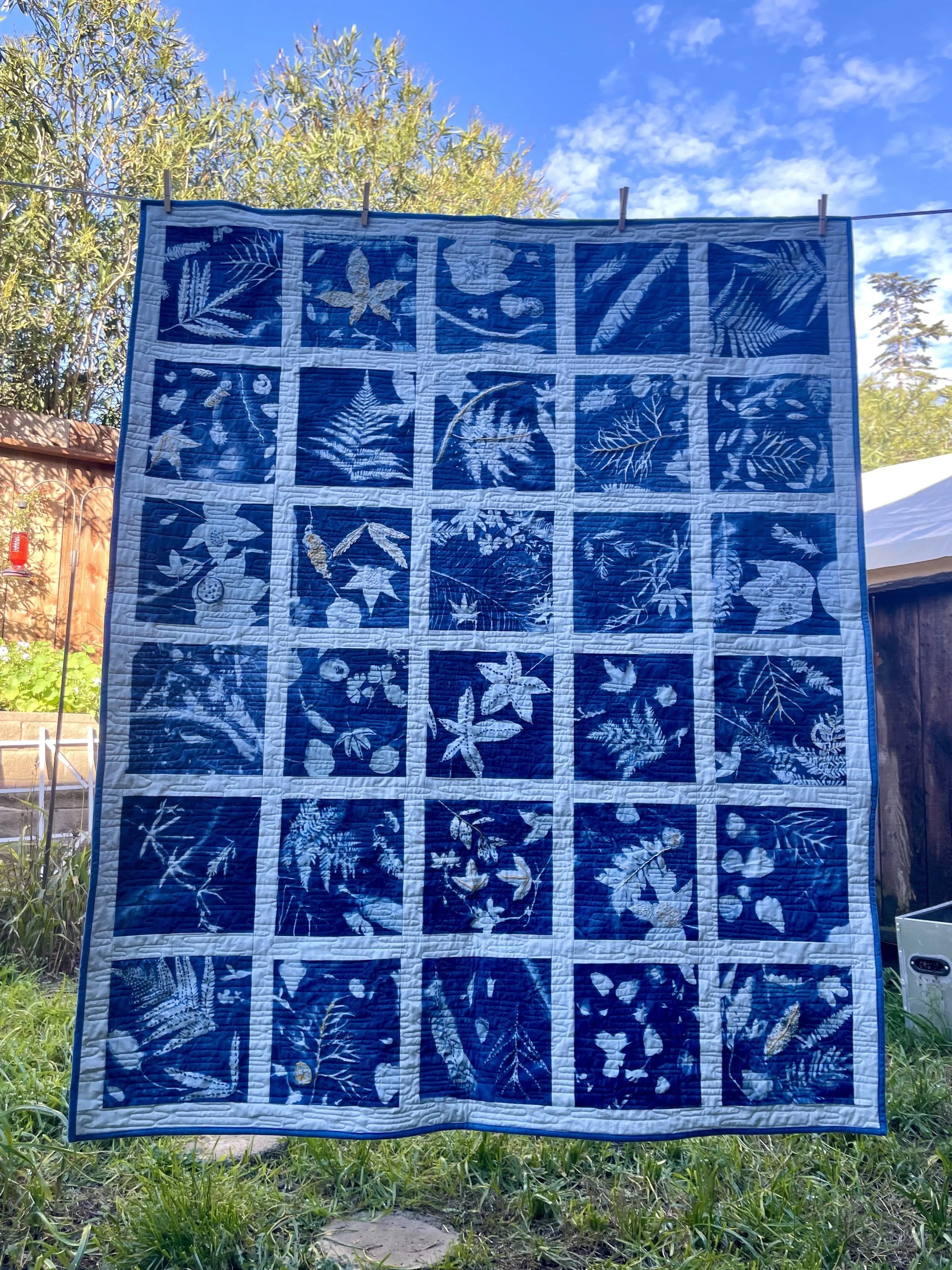 A quilt with blue and white botanical designs hanging outdoors on a clothesline against a sky with some clouds and trees in the background.