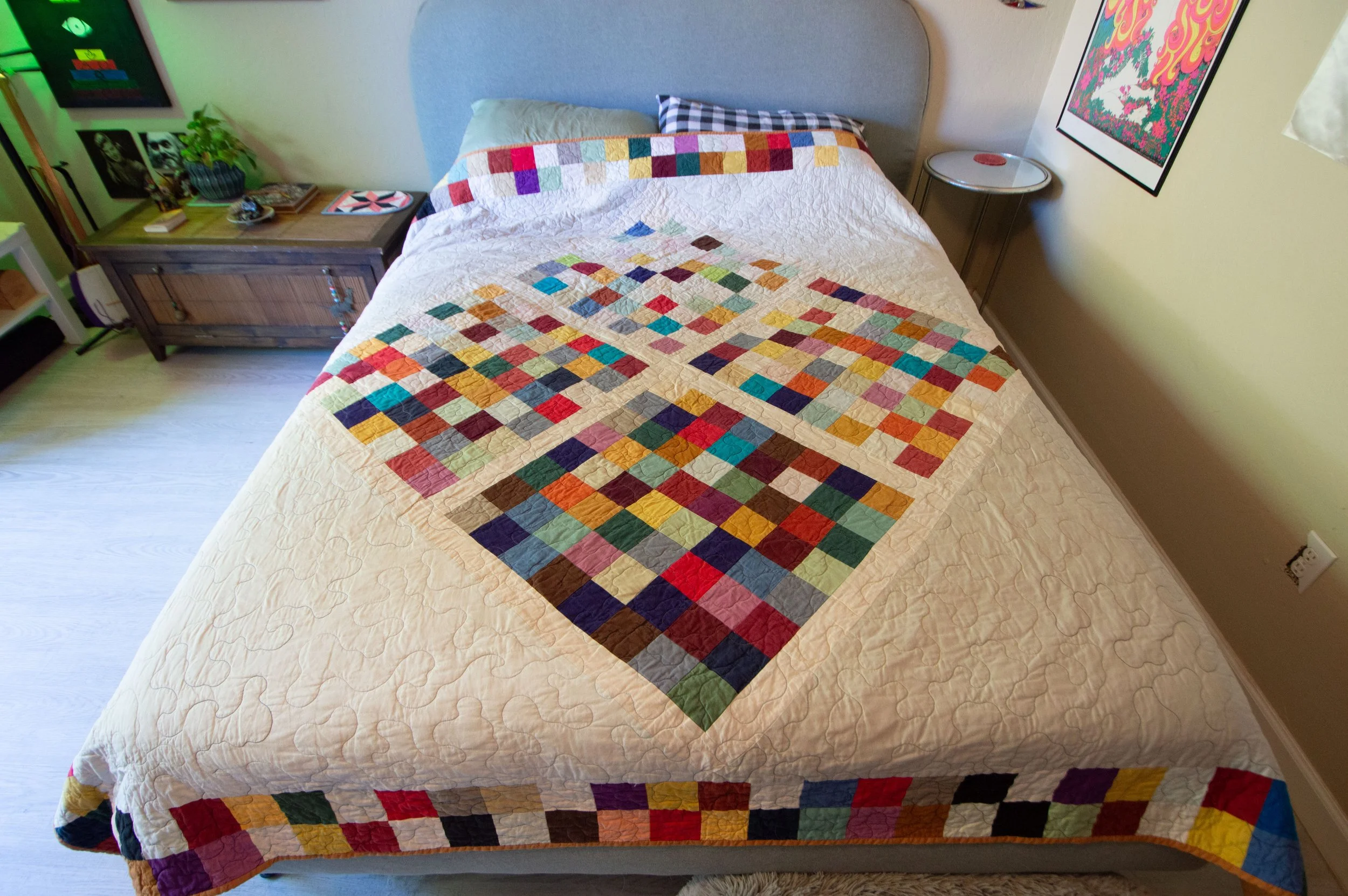 A neatly made bed with a colorful quilt featuring squares of various colors and patterns, positioned against a wall with artwork. There are two pillows, one plain and one checkered, on the bed.