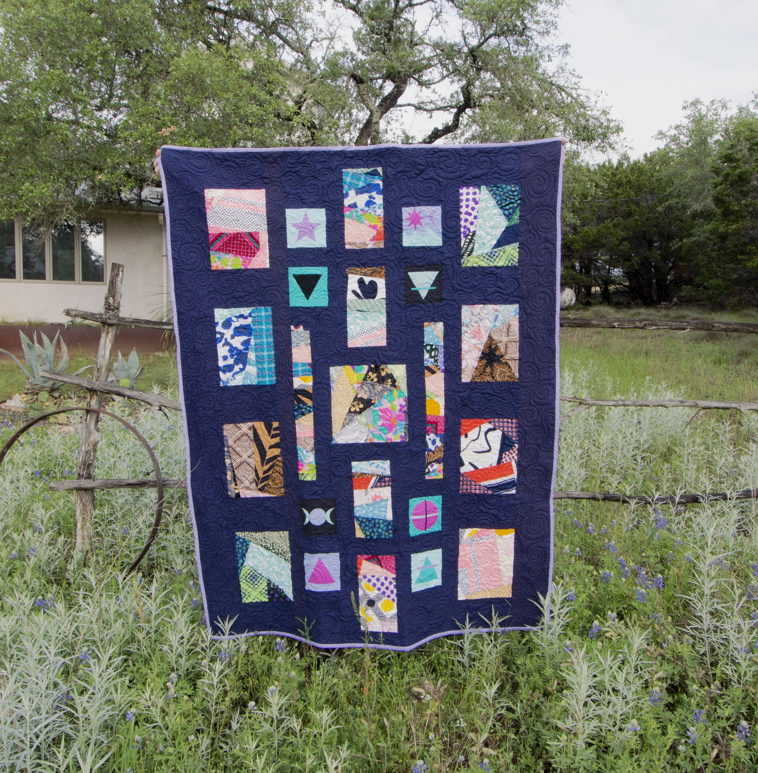 A colorful quilt with various geometric and abstract patterns hangs on a wooden fence outdoors, surrounded by green grass and plants, with trees and a building in the background.