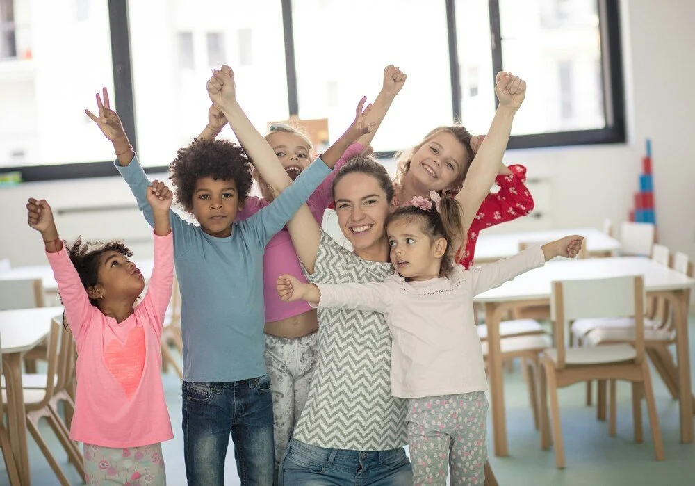 Five smiling children standing together in a classroom with their arms around each other, reflecting friendship, belonging and a welcoming school community.