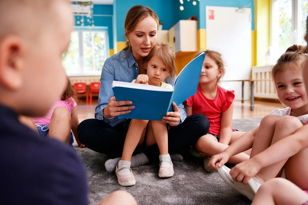 Teacher reading a book with young children seated in a classroom circle, reflecting early learning, care and a welcoming school environment.