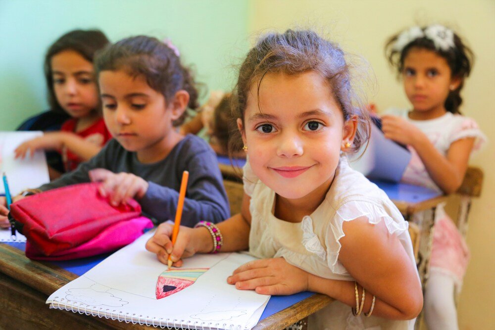 Young girl smiling at her desk while drawing in a classroom with other children, reflecting early learning, creativity and a welcoming school environment.