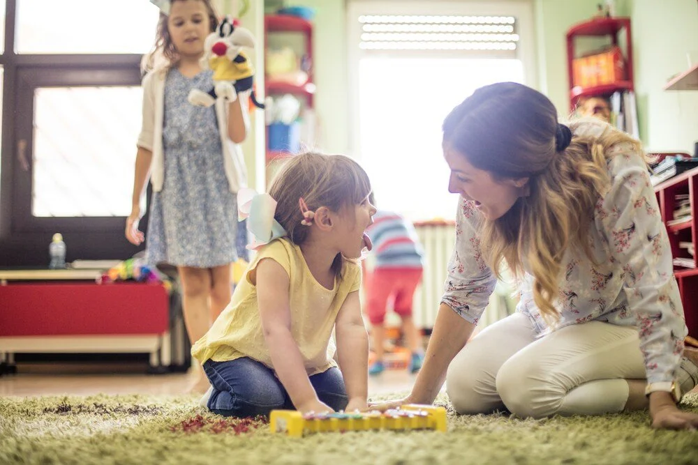 Teacher kneeling and smiling with a young child during a play-based classroom activity, with other children in the background.
