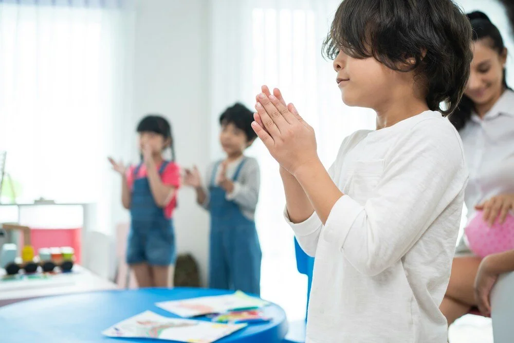 Young boy standing with hands clasped in prayer in a classroom, with classmates and a teacher in the background.