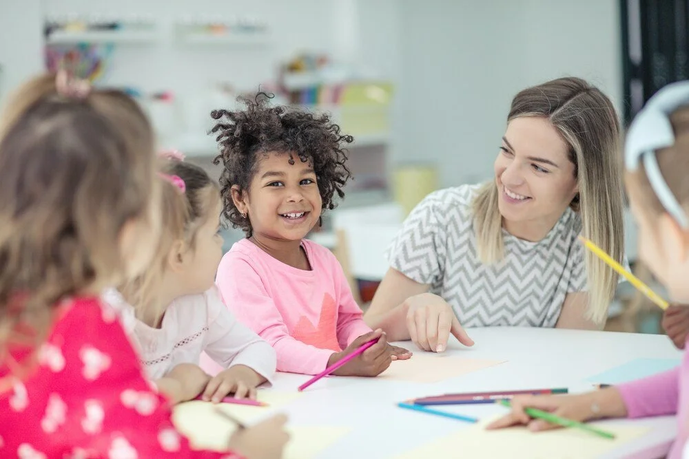 Young teacher smiling with preschool children around a classroom table during a creative learning activity.