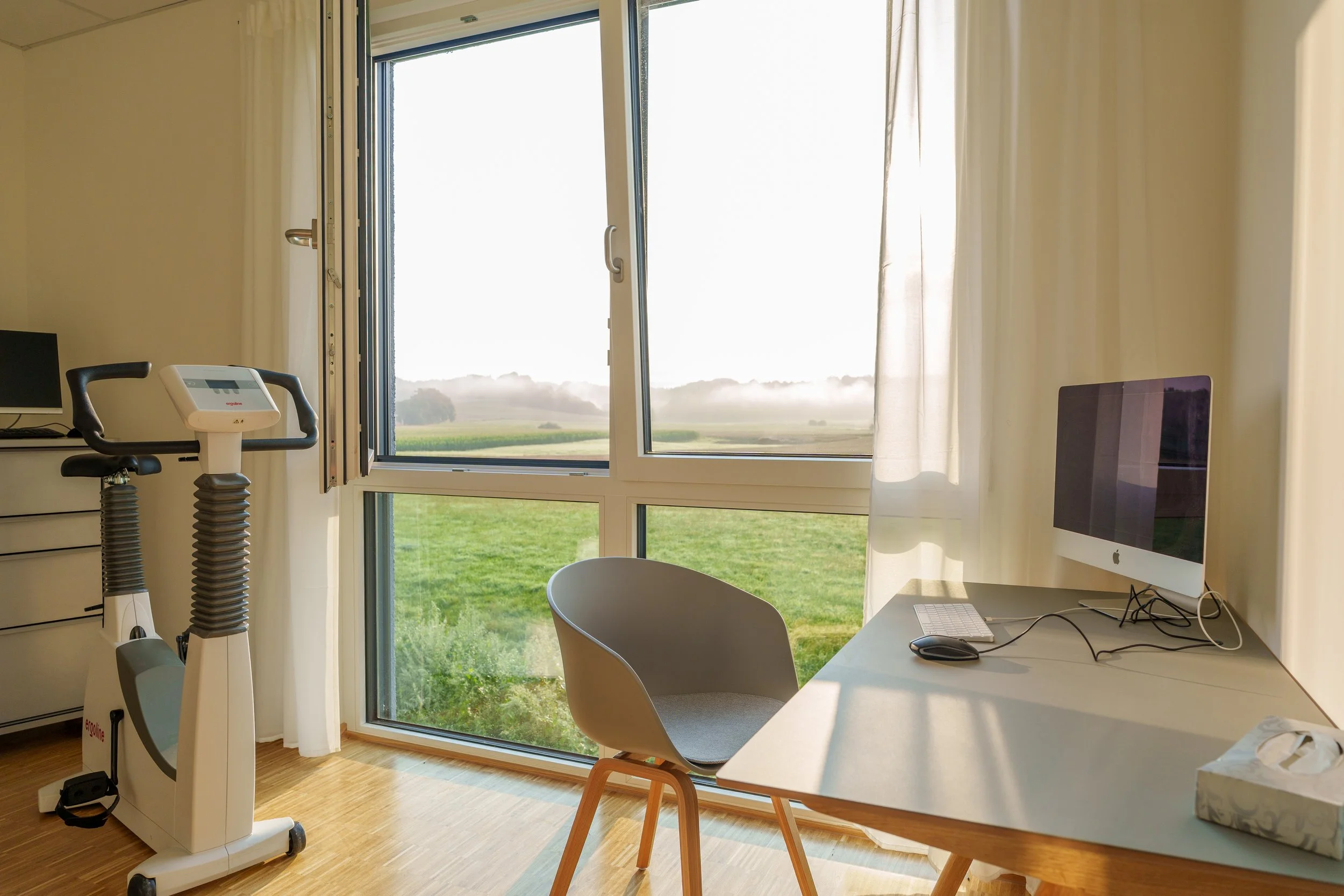Ein Büro mit Computer, Schreibtisch, Stuhl, Fahhradergometer und Blick nach draußen durch große Fenster auf eine grüne Landschaft bei Sonnenaufgang.