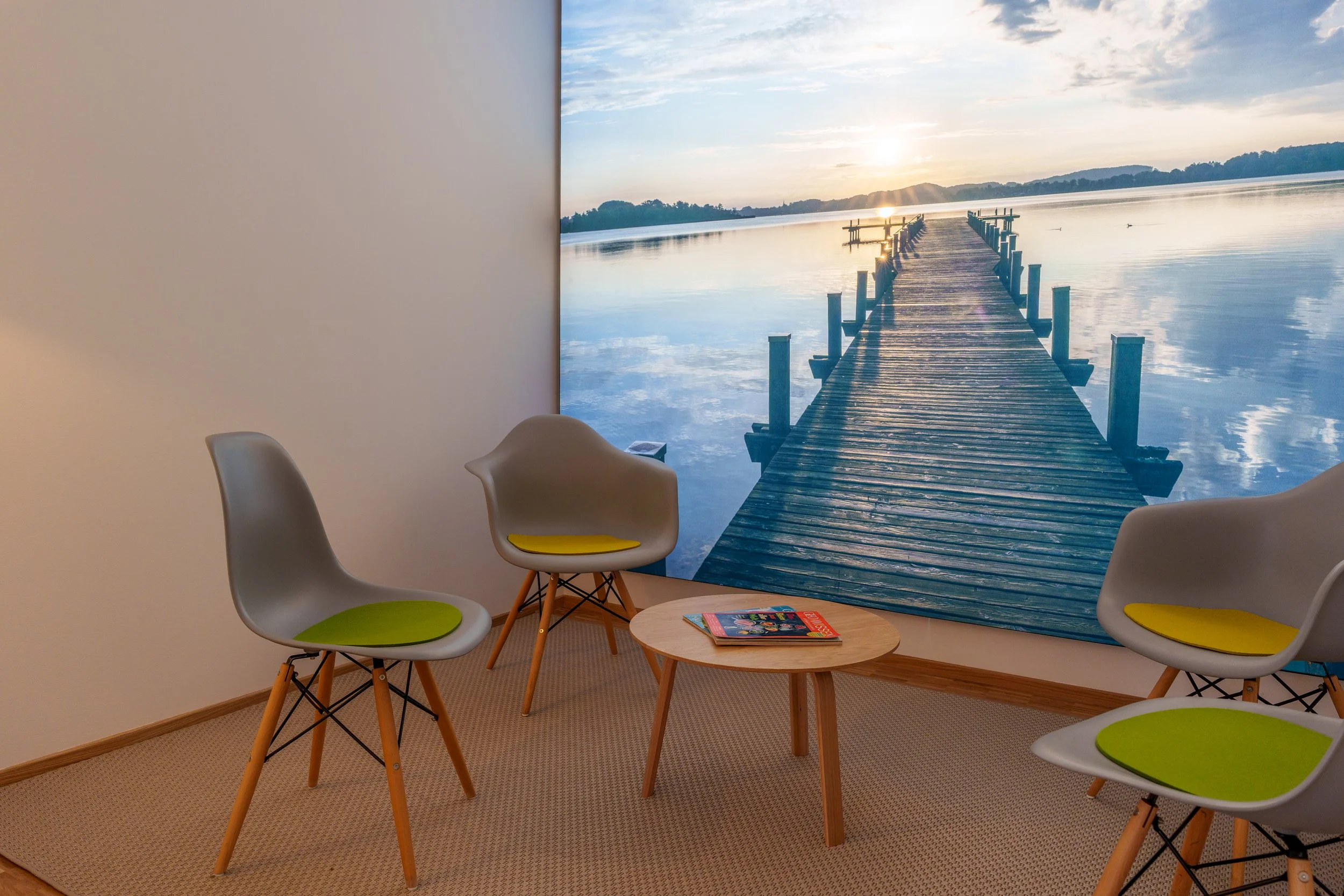 Wartenbereich mit vier Stühlen und einem kleinen Tisch vor einer Wand mit Fototapete eines Steg auf einem See bei Sonnenuntergang.