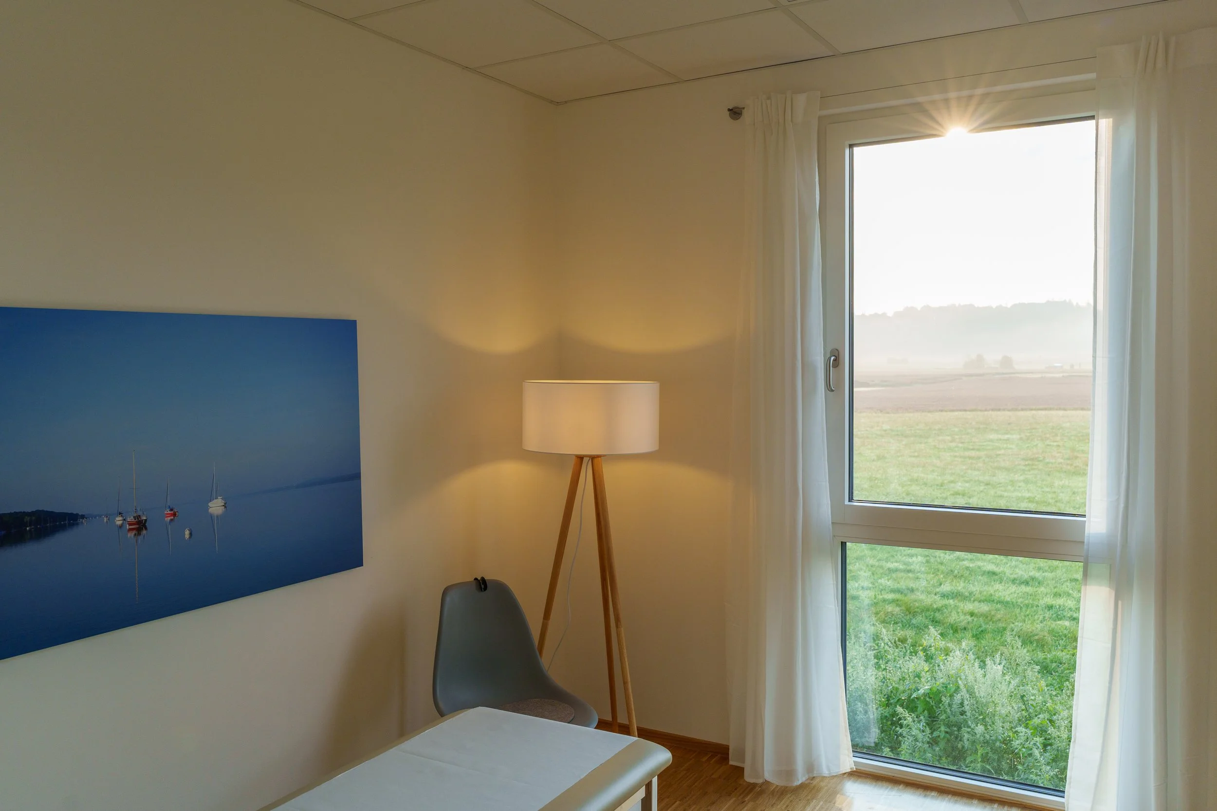 Ein komfortables Schlafzimmer mit einem Fenster, das den Blick ins Grüne freigibt, einem Nachtisch, einem Stuhl, einer Stehlampe und einem Wandbild von Booten auf Wasser.
