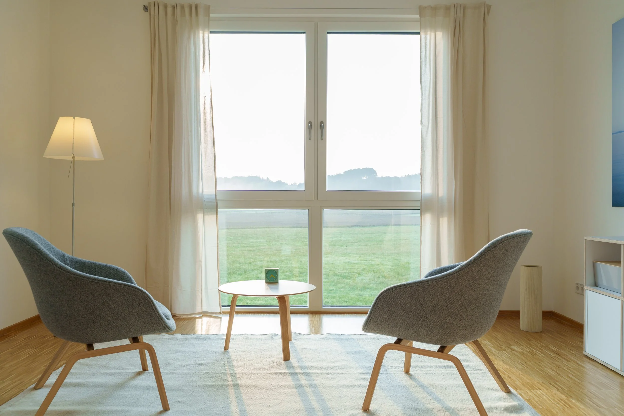 Therapieraum mit zwei grauen Sesseln, einem kleinen runden Tisch, einem großen Fenster mit weißen Vorhängen, Holzboden und auf dem Tisch steht eine kleine Uhr.