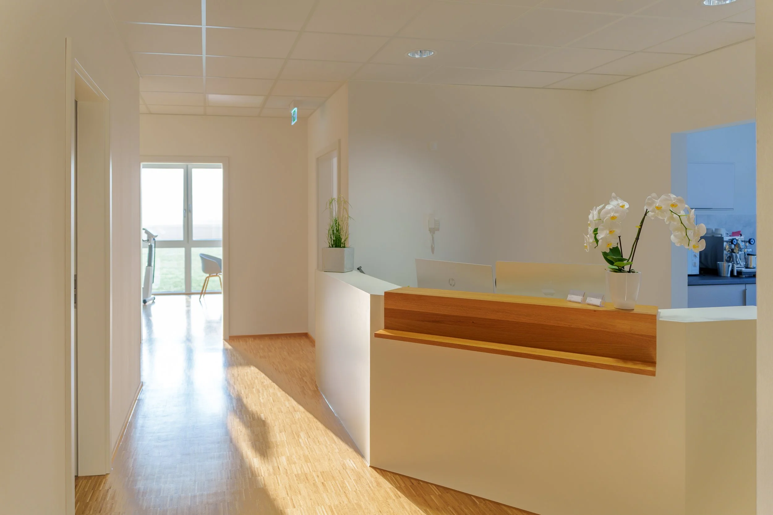 Heller Empfangstresen mit weißen und holzfarbenen Elementen, eine Topfpflanze mit weißen Orchideen, Blick auf einen Flur mit Tür und Fenster, im Hintergrund ein Raum mit Kaffeeautomaten.