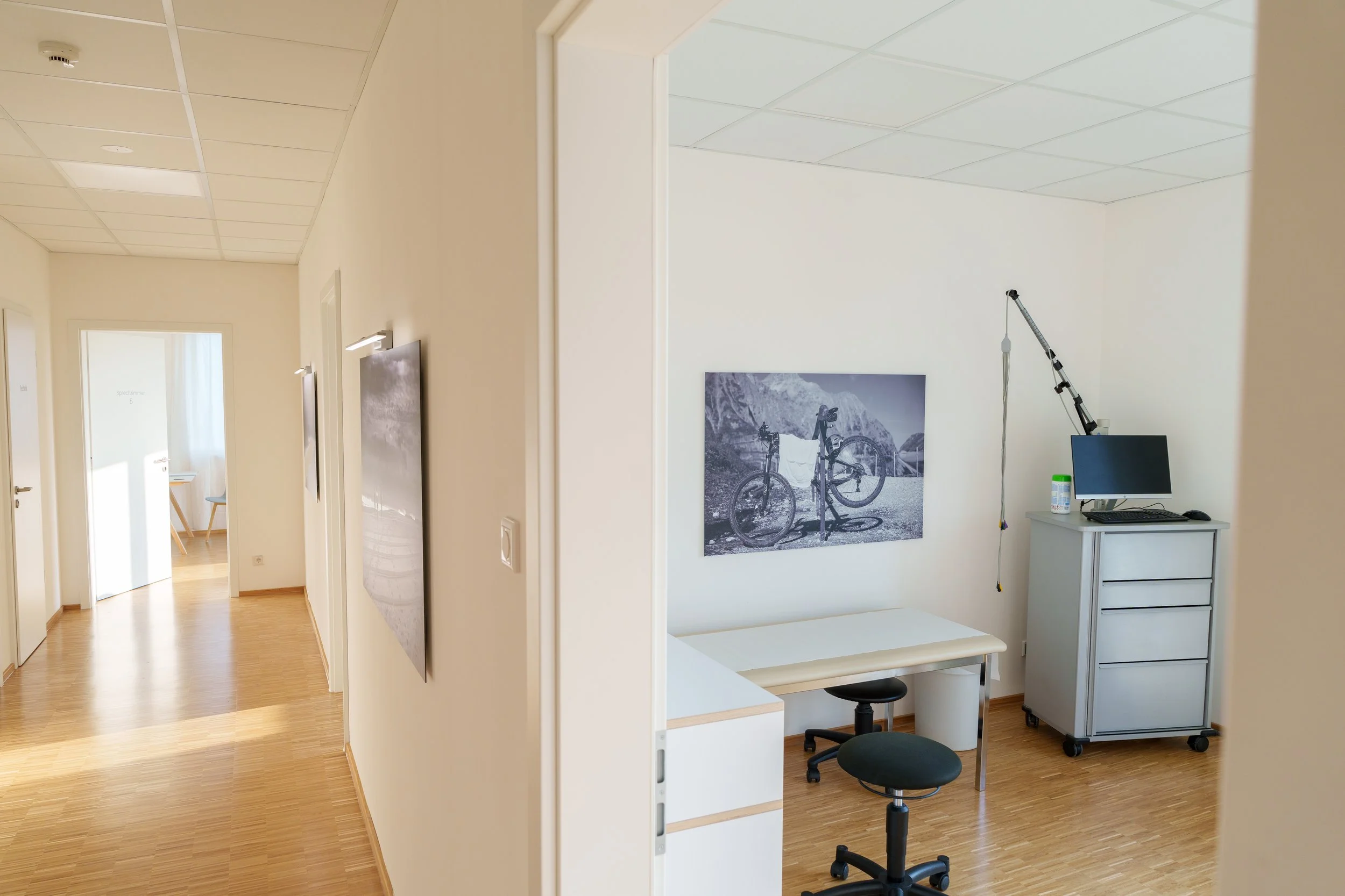 Innenansicht eines medizinischen oder therapeutischen Raums mit einem Untersuchungstisch, Stühlen, einem Bildschirm und medizinischer Ausrüstung, mit einem Fahrradbild an der Wand.