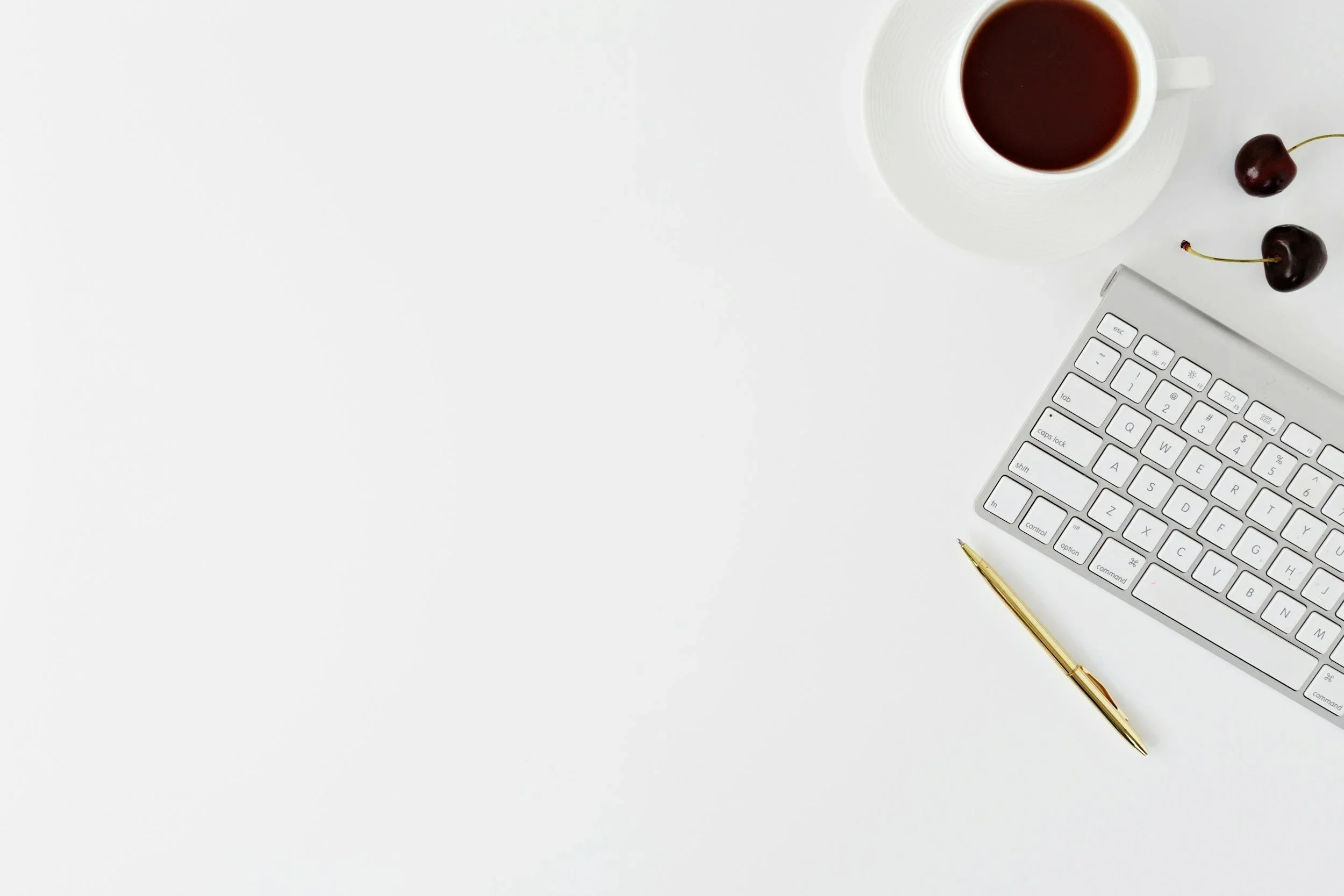 A white workspace with a computer keyboard, a gold pen, a cup of coffee on a saucer, and two cherries on a white surface.