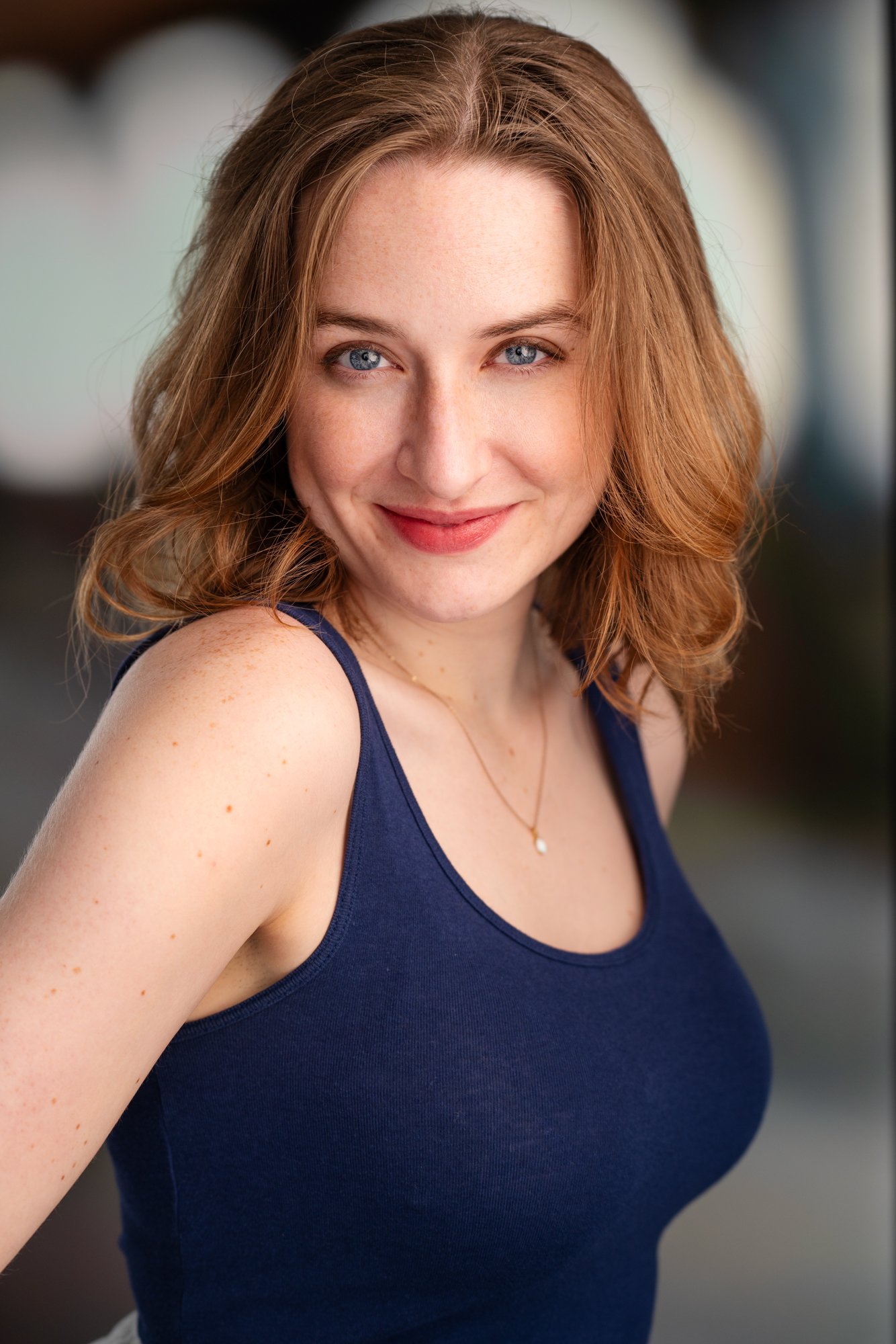 A woman with shoulder-length red hair, blue eyes, wearing a navy blue tank top and a delicate necklace, smiling warmly at the camera.