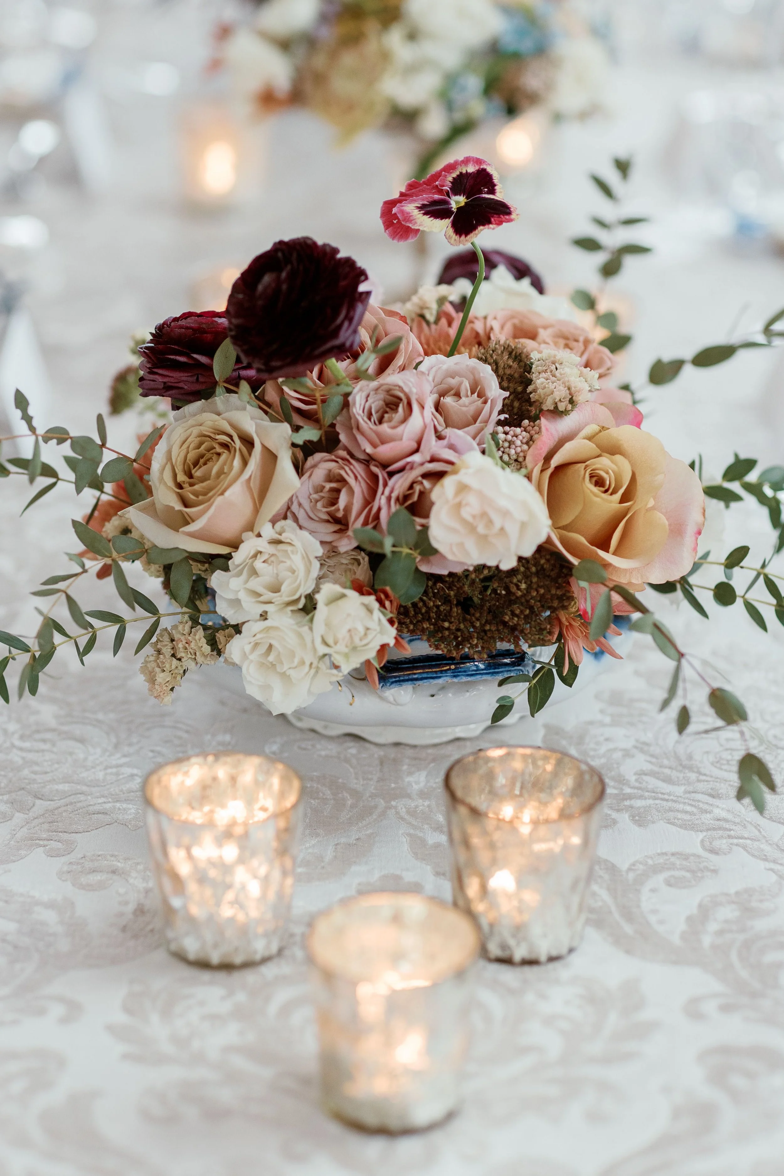 A floral centerpiece with pink, white, and purple flowers on a table with white lace tablecloth and lit candles in decorative holders.