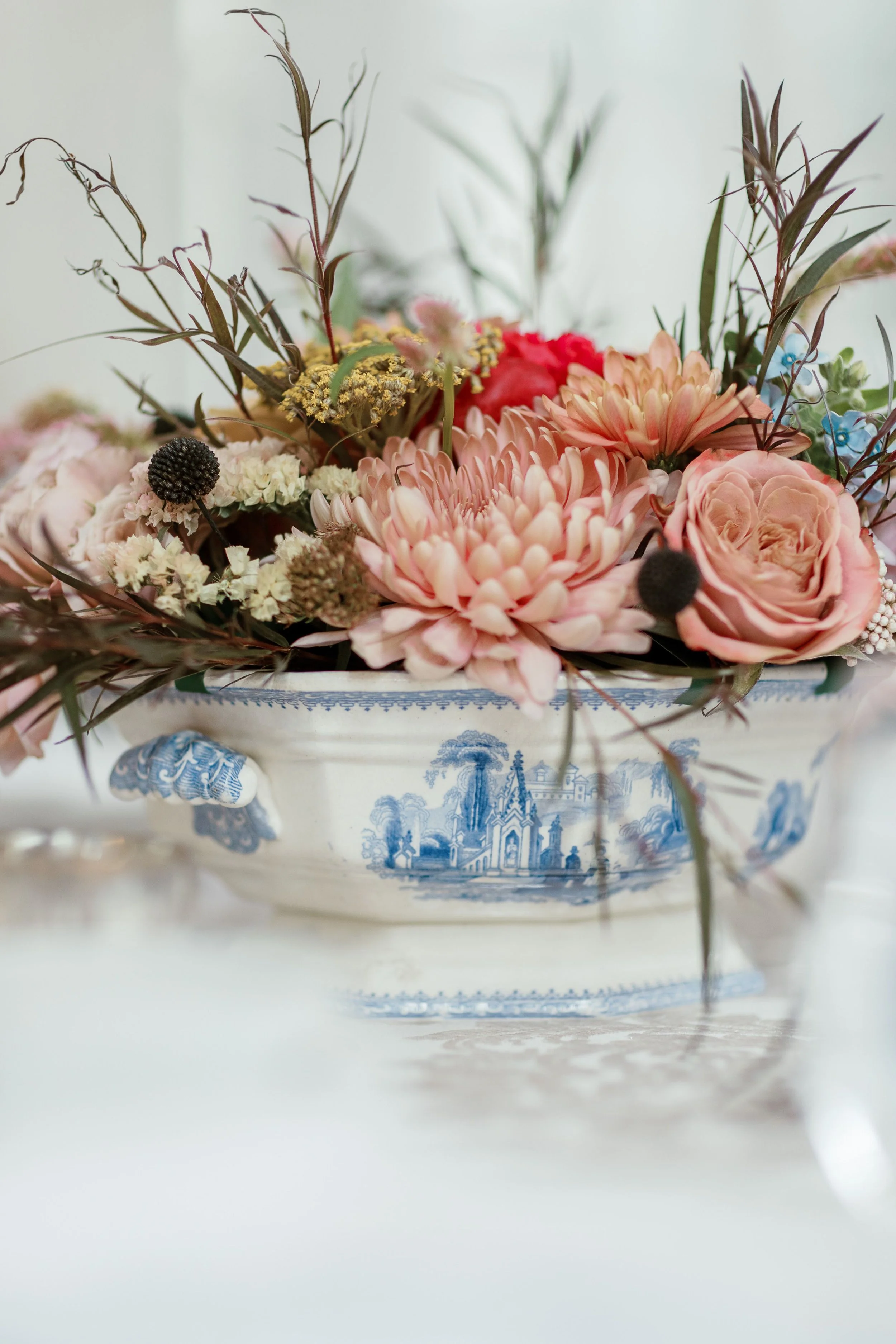 A floral arrangement in a white bowl with blue decorative scenes, containing pink and peach flowers along with dried greenery and small accents.