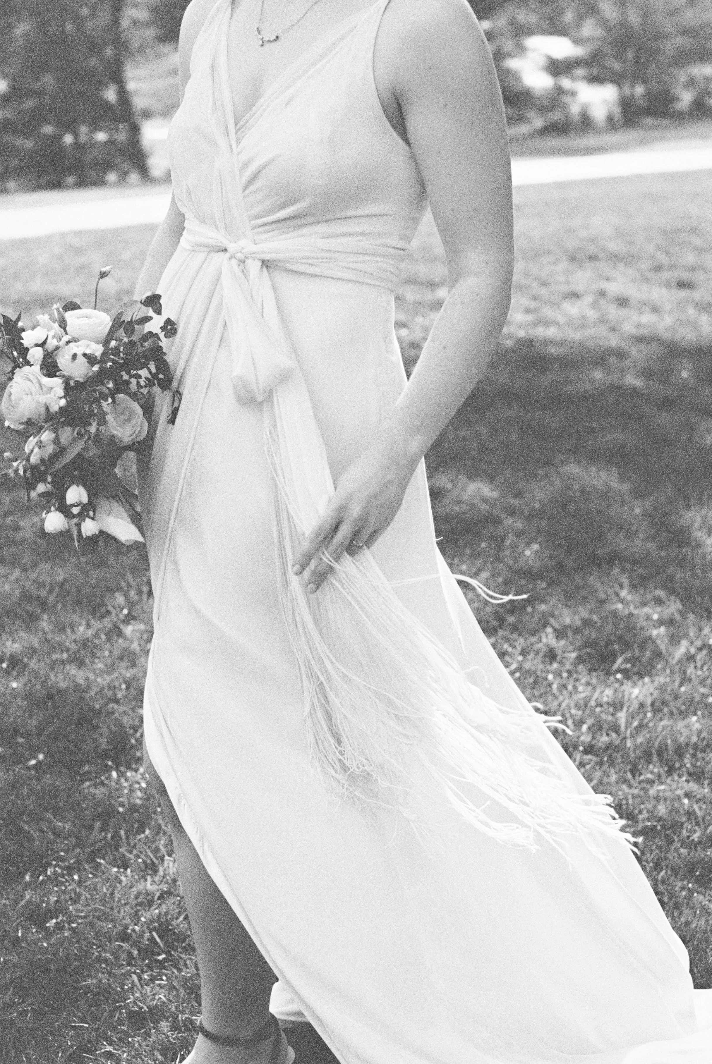 A woman in a flowing, sleeveless, light-colored dress holding a bouquet of flowers, standing on grass with trees or bushes in the background.