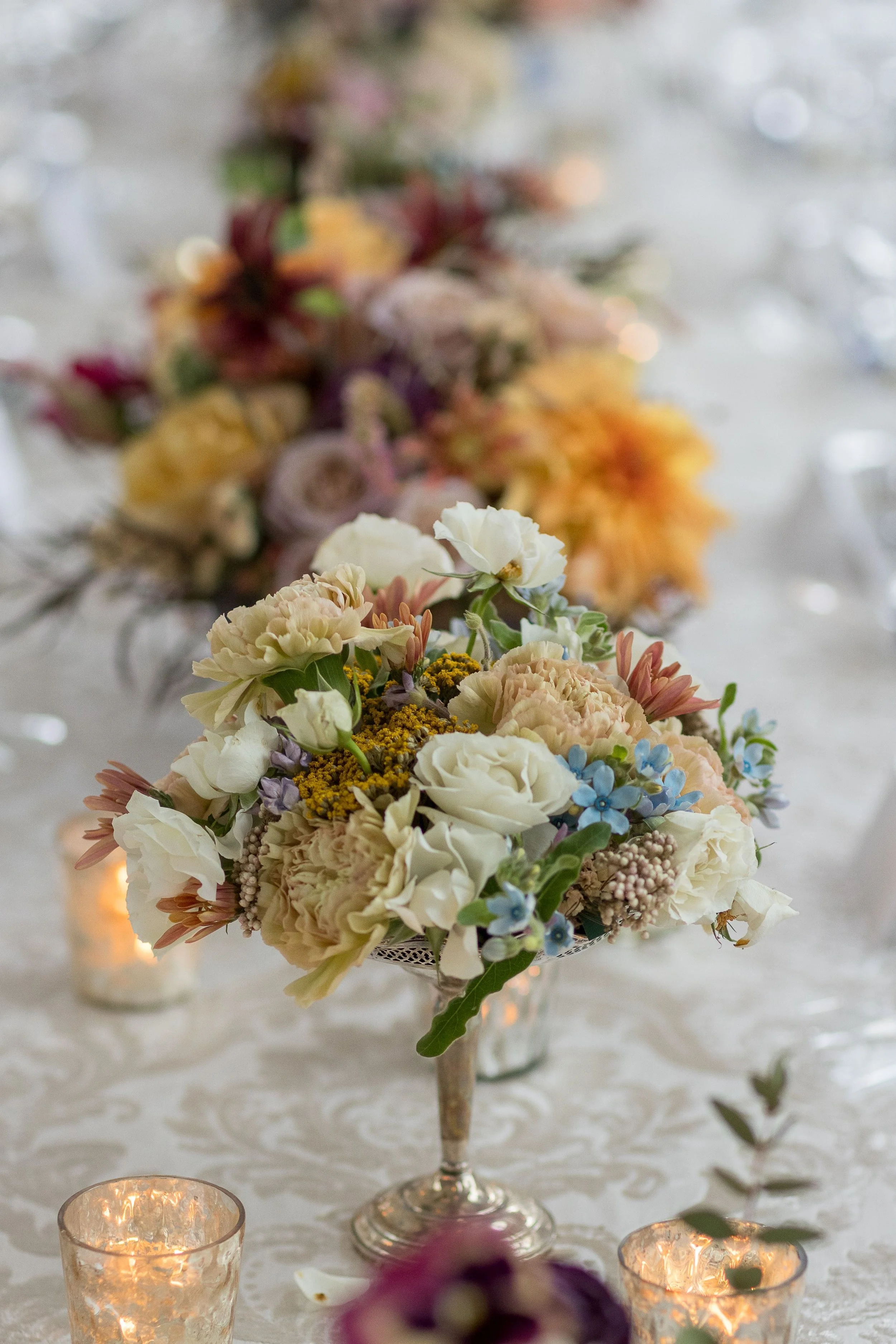 Floral centerpiece with white, pink, and purple flowers on a table surrounded by candles at an event.