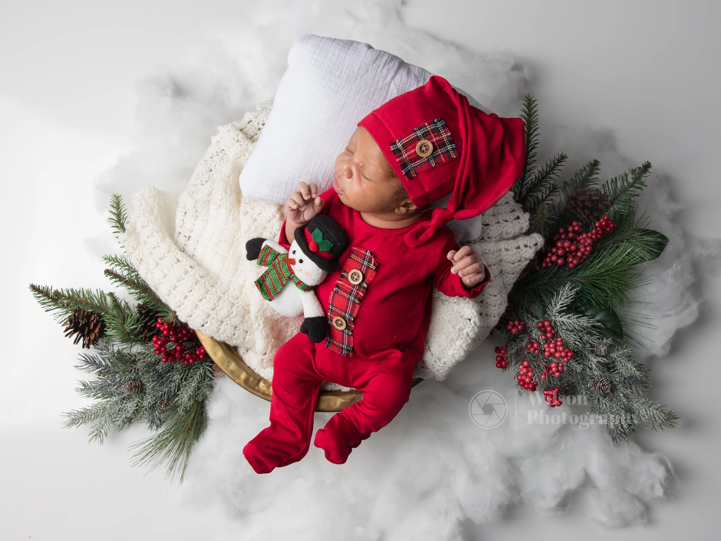Newborn baby dressed in a festive red outfit with plaid details and wooden buttons. The baby is lying on a soft, white blanket inside a circular basket surrounded by holiday-themed decorations such as pine branches, red berries, and a plush snowman t