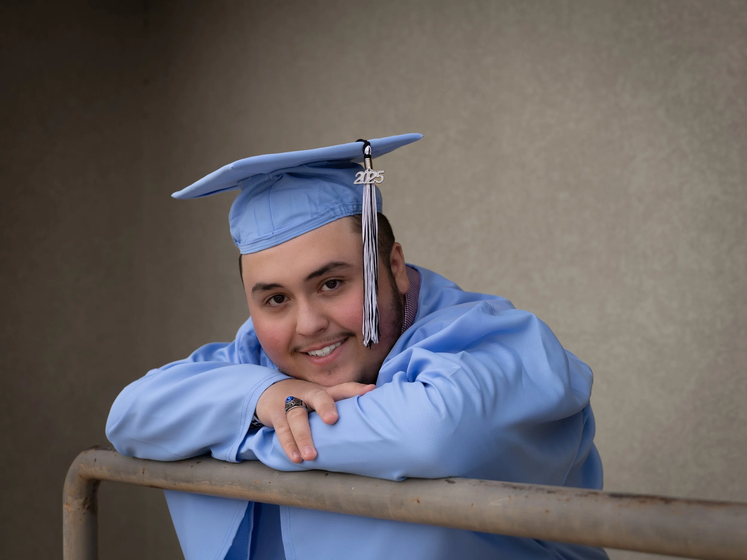 A young man wearing a light blue graduation gown with a 2025 tassel on the hat and blue senior ring.