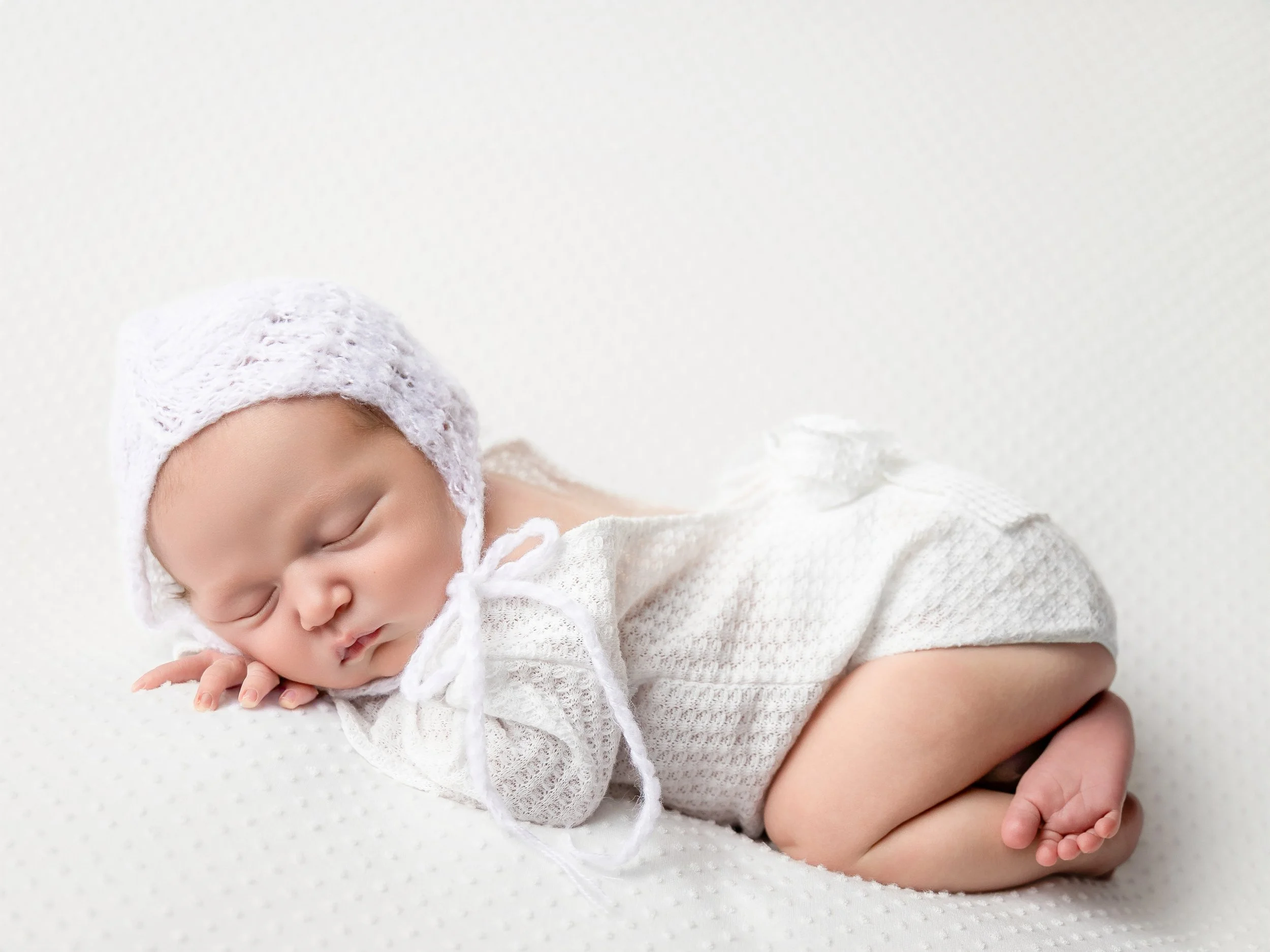 A Newborn girl wearing a white bonnet and a white outfit laying on her belly with her hand posed under her head and her feet tucked under her bottom.