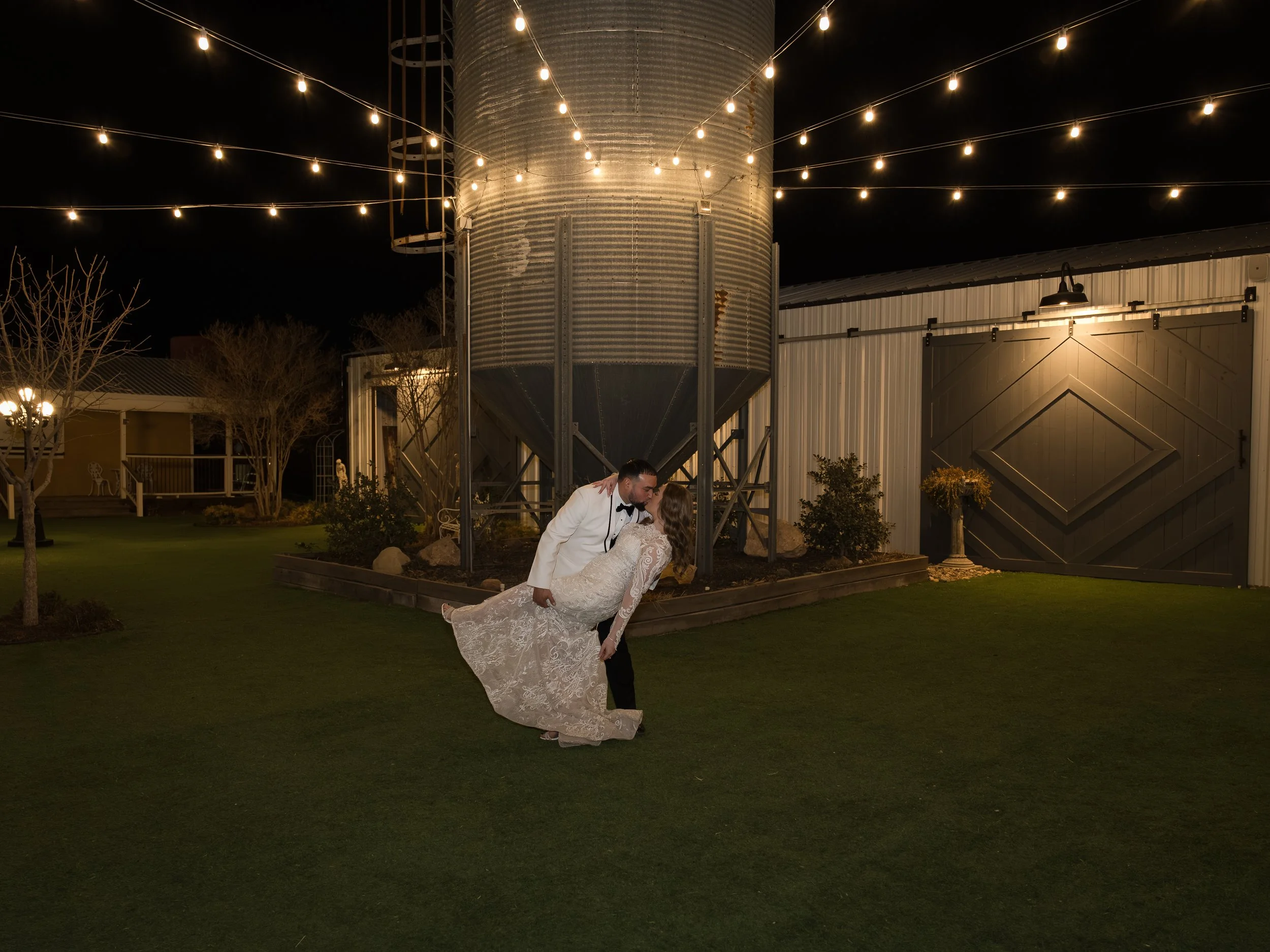 A romantic scene featuring a couple at a wedding. The groom, in a white jacket and black pants, dips the bride, who is wearing a long white lace gown, for a kiss. They are outdoors at night, with a metal silo decorated with string lights in the backg