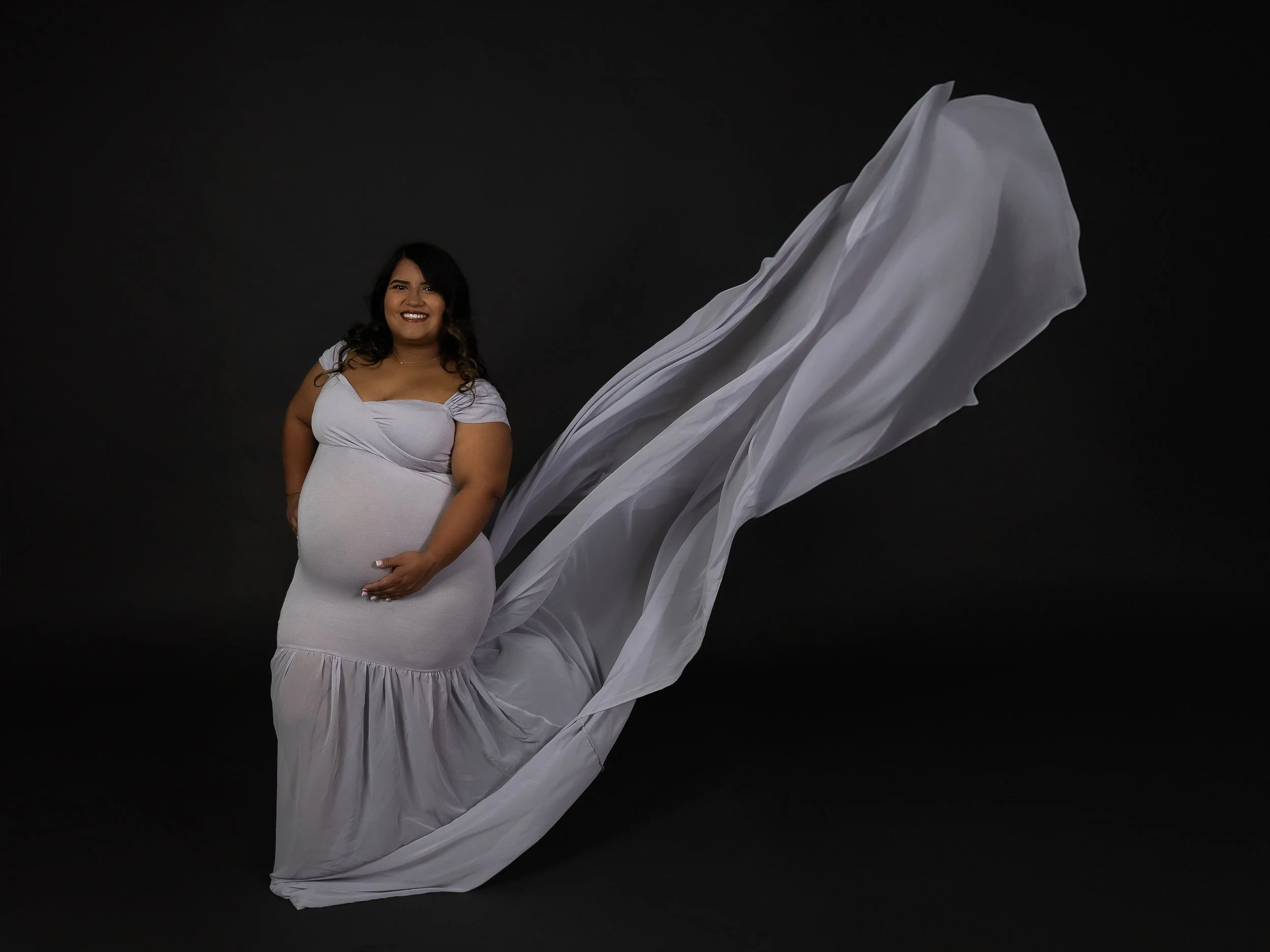 The image portrays a pregnant woman in a gray, flowing gown with off-the-shoulder sleeves, set against a dark background. Her gown drapes elegantly behind her, adding to the dramatic effect. She smiles softly while gently holding her baby bump.