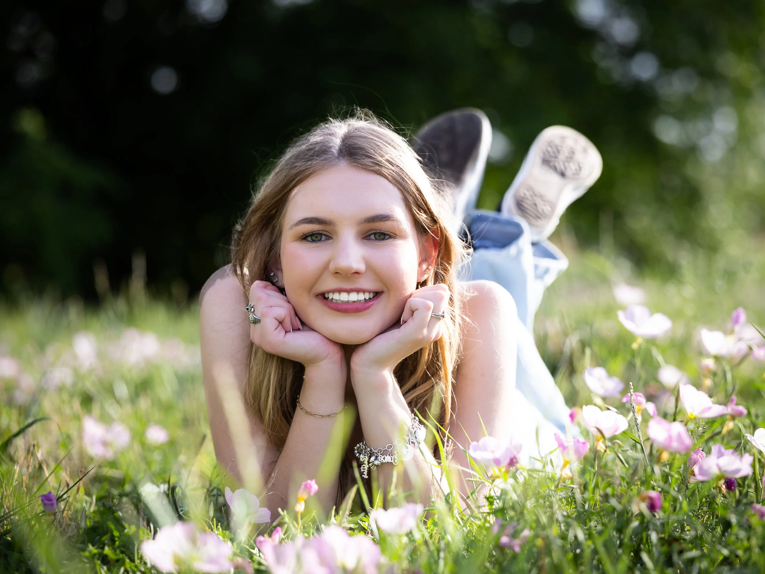 A young woman posing while laying on grass with wildflowers wearing jeans and has her shoes showing.